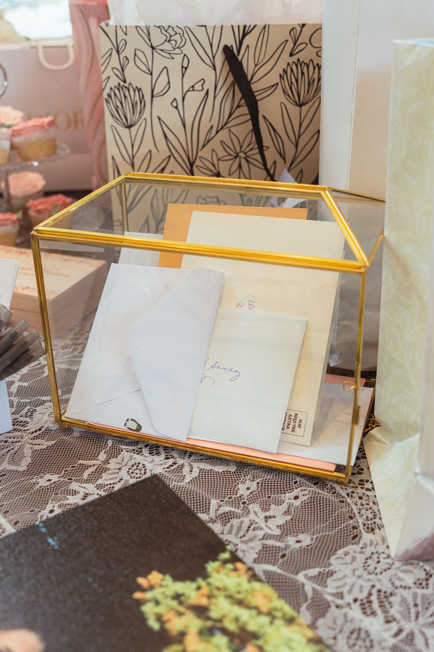 Gold glass display box filled with envelopes and cards on lace fabric, surrounded by floral gift bags.