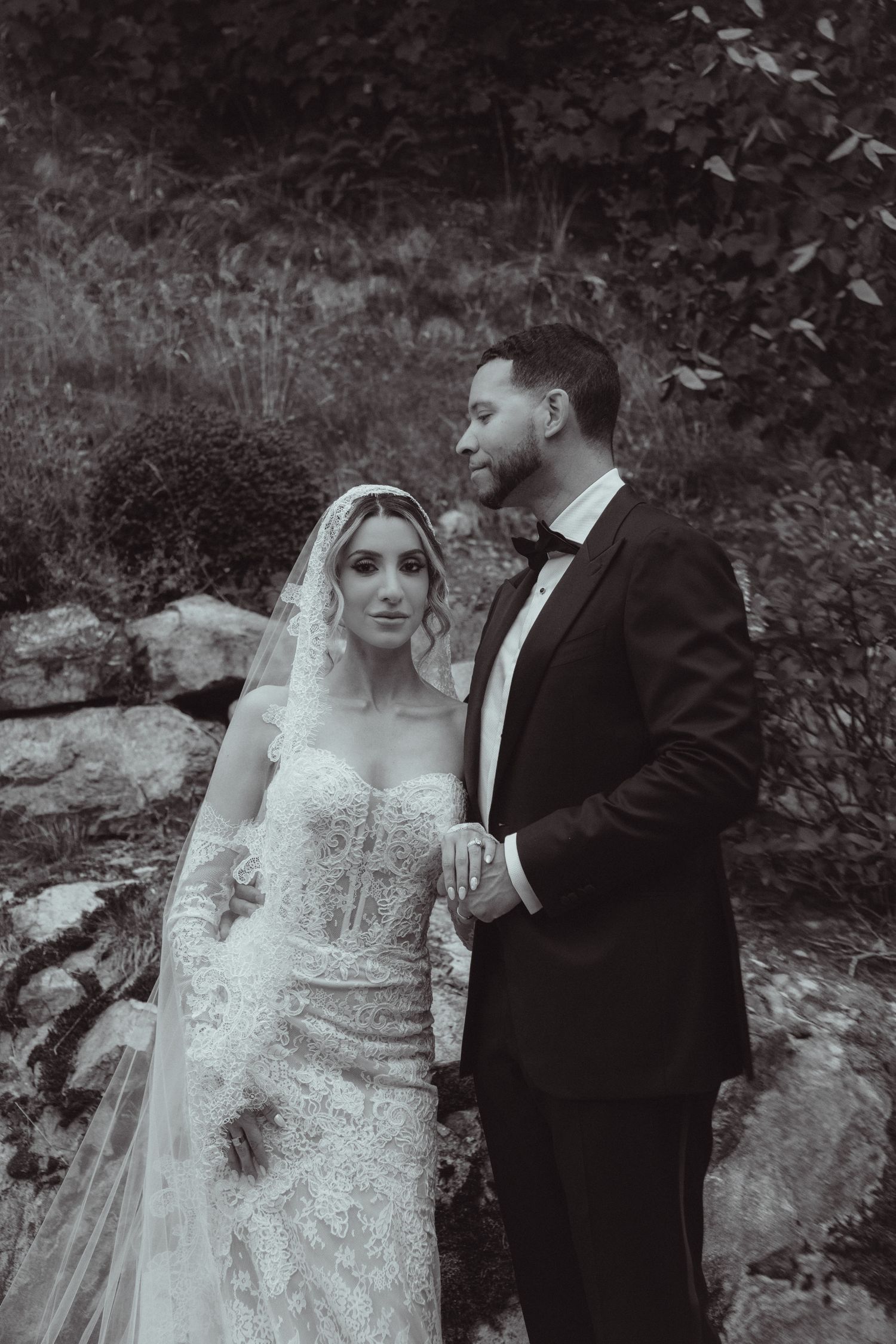 Black and white wedding photo of bride in lace gown with veil and groom in tuxedo posing outdoors among rocks and foliage.