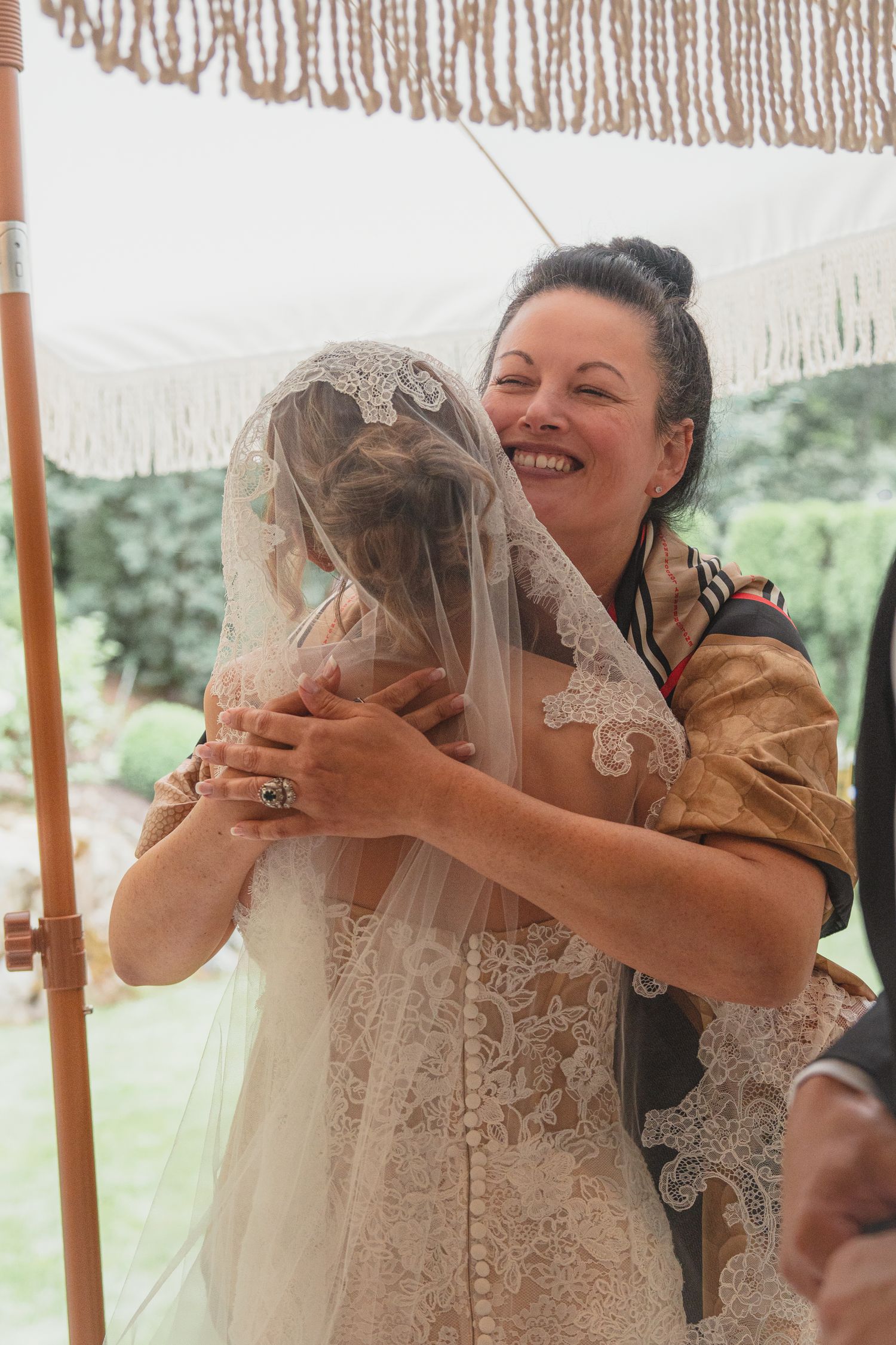 A joyful woman in golden attire hugs a bride in a lace veil during an outdoor wedding ceremony.