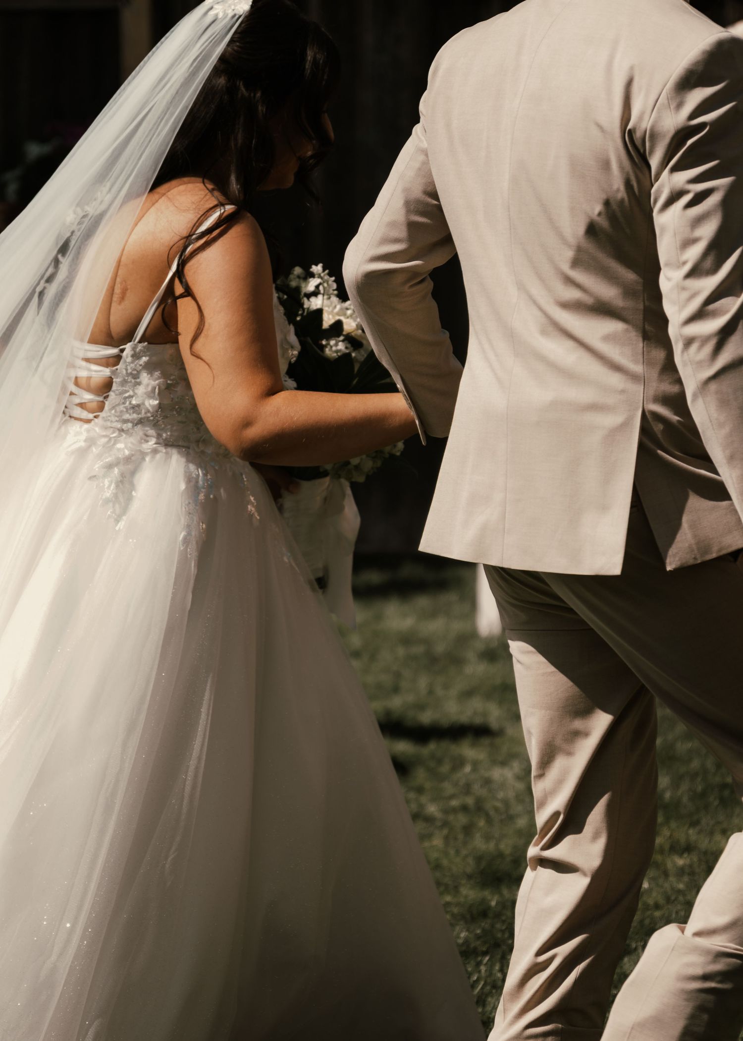 Bride in white lace gown with veil and groom in tan suit walking together outdoors on their wedding day.