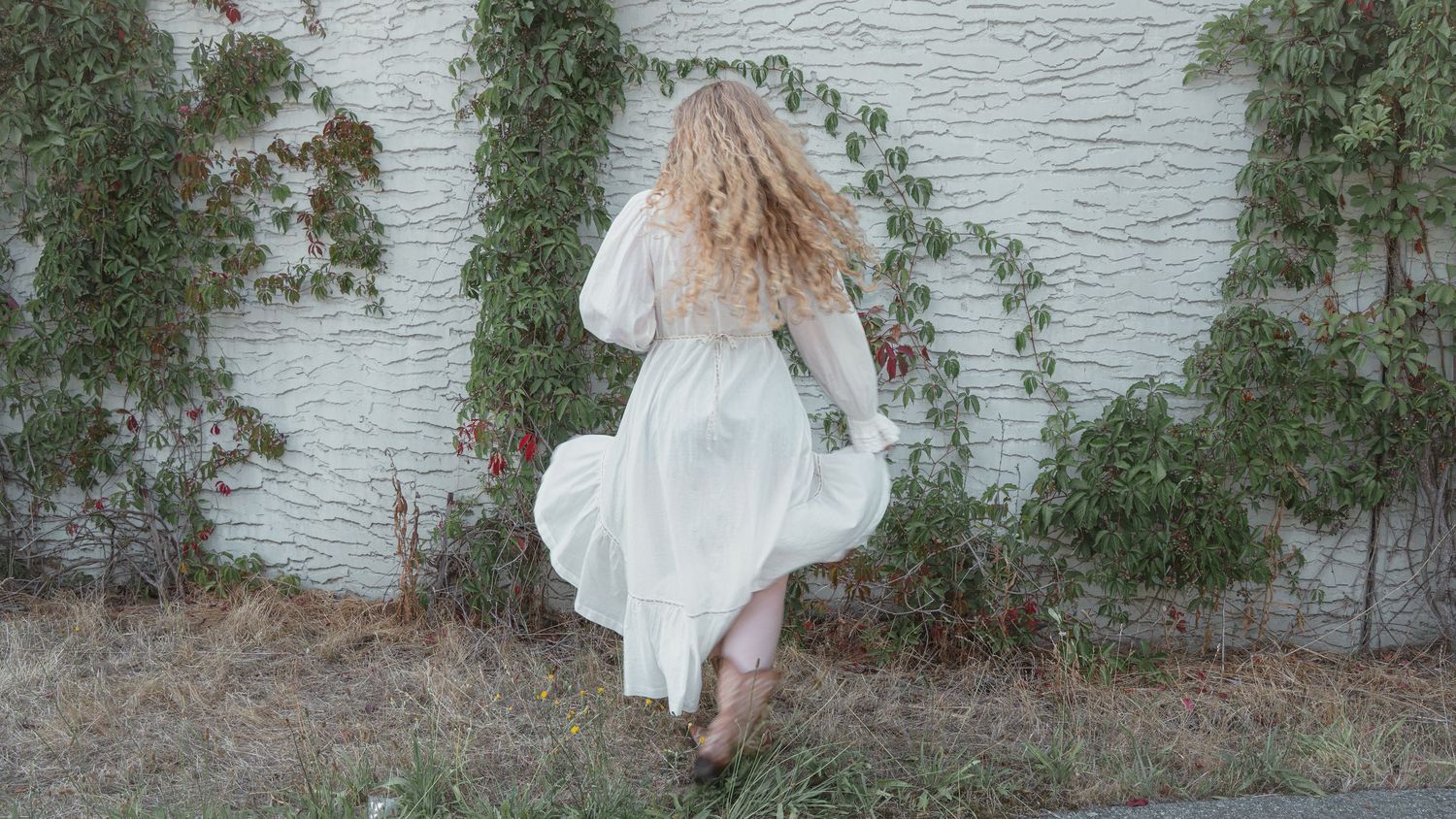 A woman with curly blonde hair in a flowing white dress stands near an ivy-covered white brick wall outdoors.