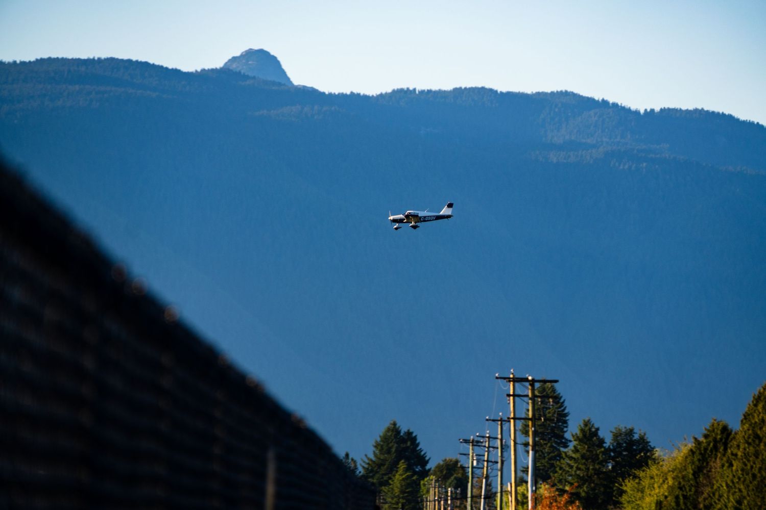A small plane flies over power lines against a backdrop of a large forested mountain under a clear blue sky.