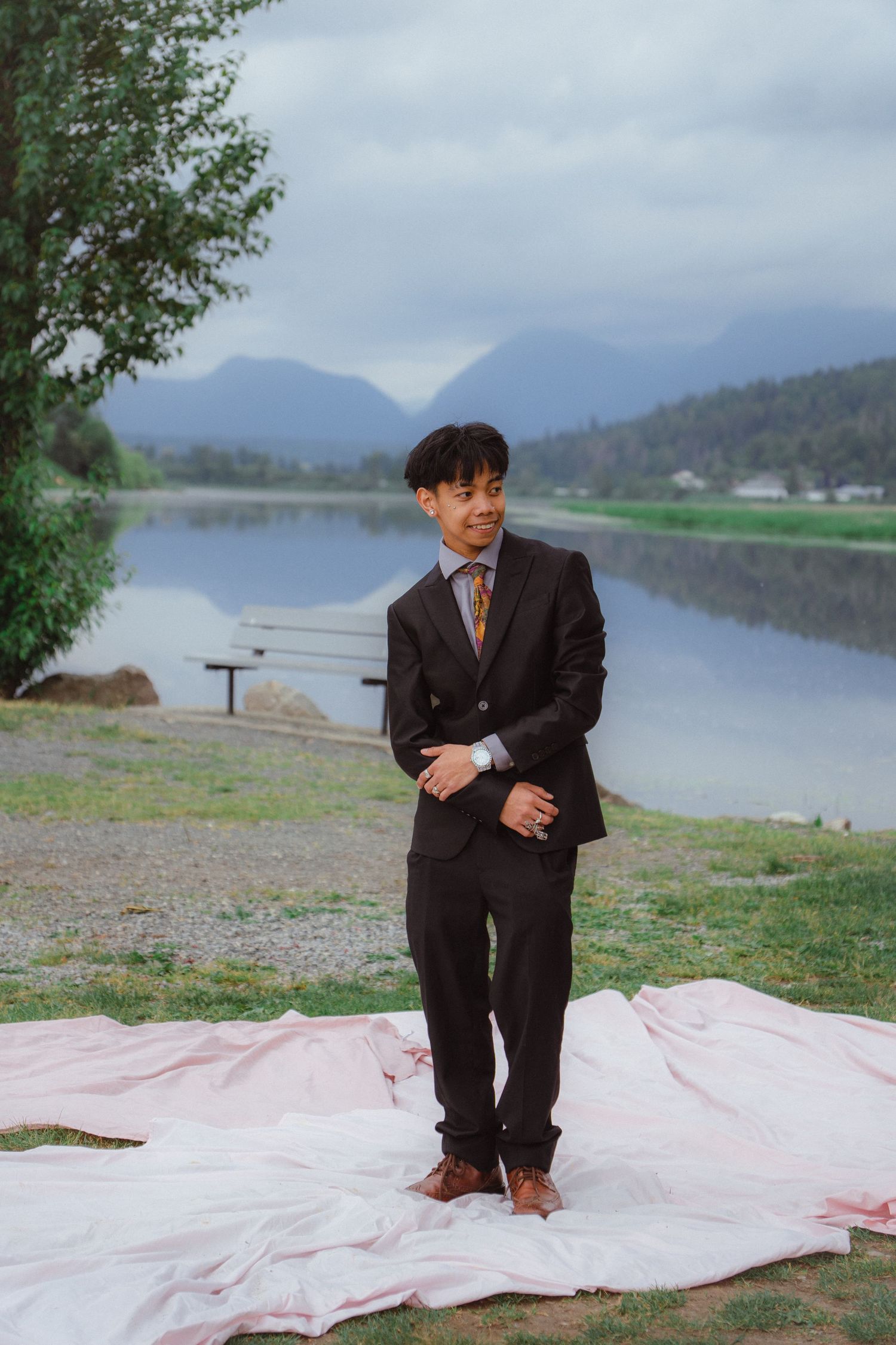 Young man in black suit stands confidently on pink blanket by a misty mountain lake.