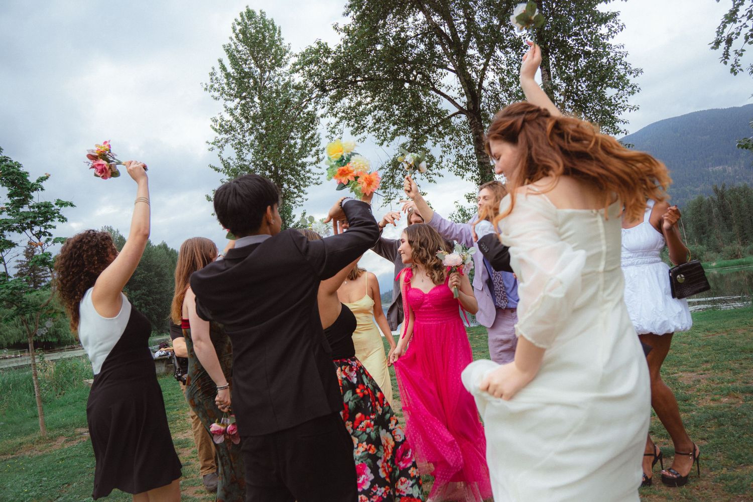 Bride tosses bouquet to guests at outdoor wedding celebration with trees and mountains in background.