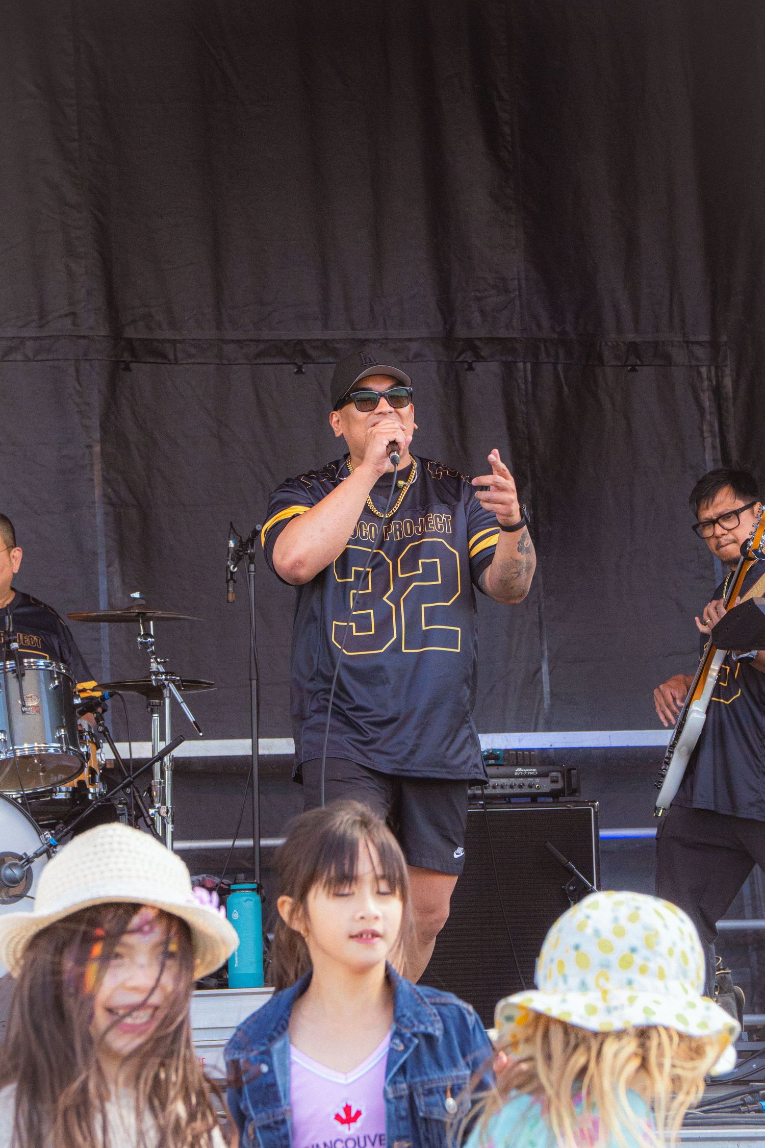 Singer in jersey #32 performs on outdoor stage while children dance in foreground at music festival.