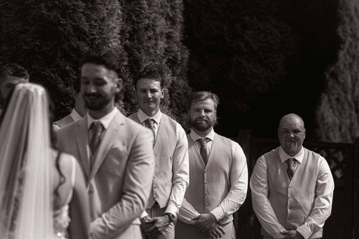 Black and white photo of groomsmen standing at outdoor wedding ceremony, smiling as bride approaches the altar.
