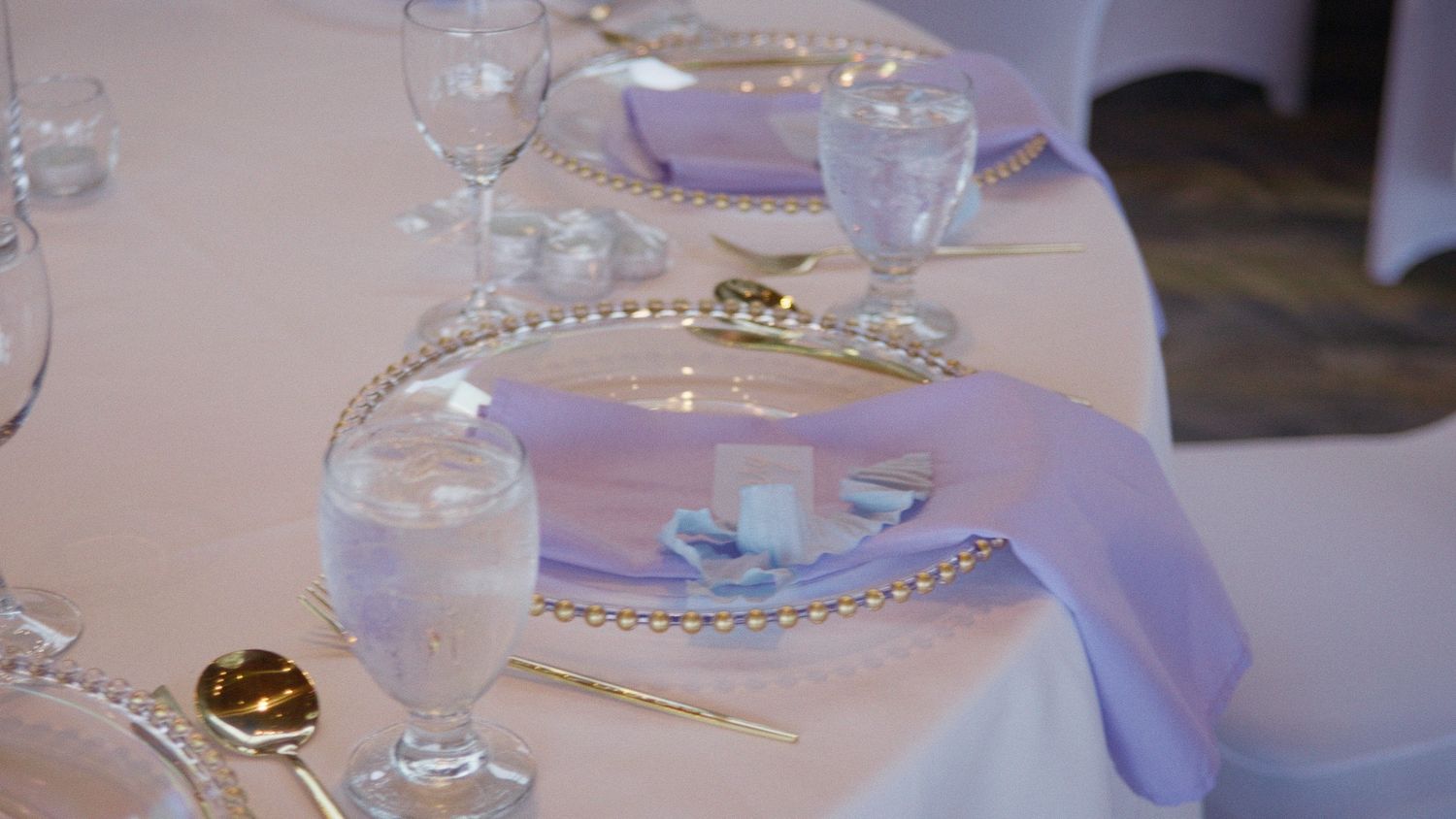 Elegant wedding table setting with gold-rimmed charger plates, crystal glasses, and lavender napkins on a white tablecloth.