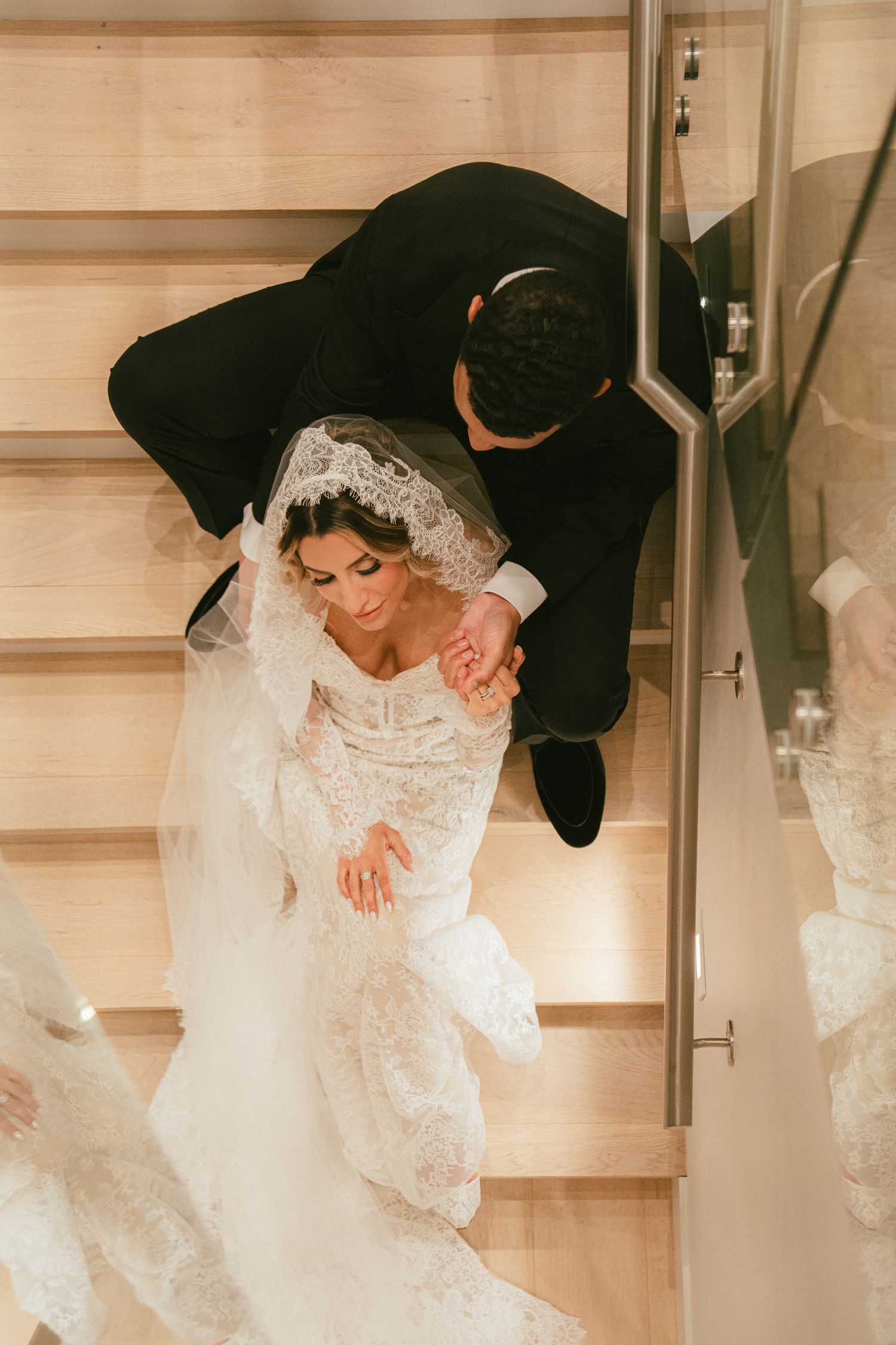 Aerial view of bride in lace gown and veil with groom in black suit descending elegant wooden staircase.
