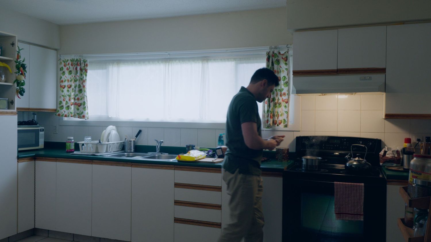 Man stands at a modern white kitchen counter preparing food near a stove with colorful curtains in background.