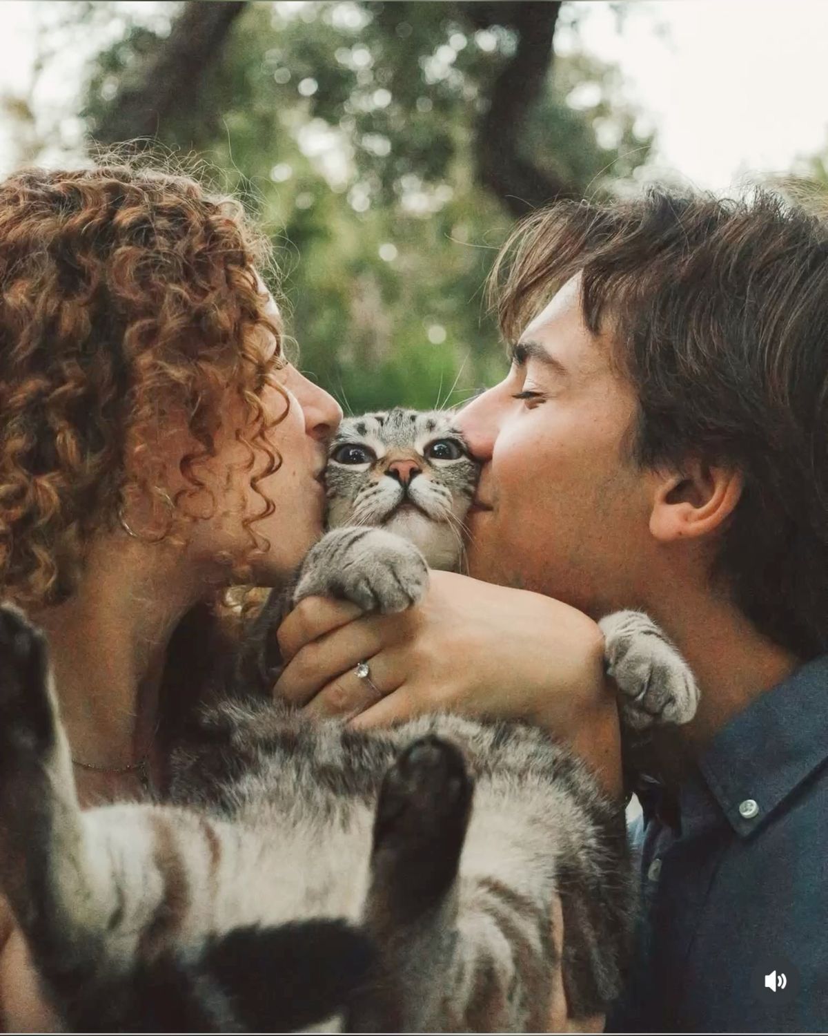 A couple with curly and straight hair kisses a gray tabby kitten on the cheeks outdoors.