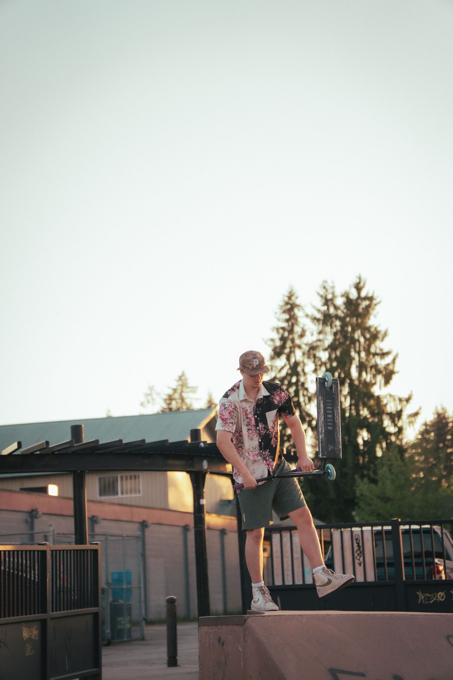 Young man in floral shirt posing with a scooter on a skate park ramp at sunset.