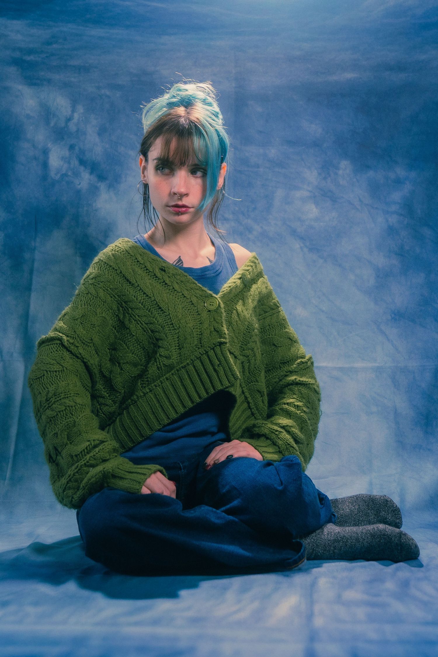 Woman with blue-dyed hair in green knit cardigan and jeans sits cross-legged on floor in blue studio.
