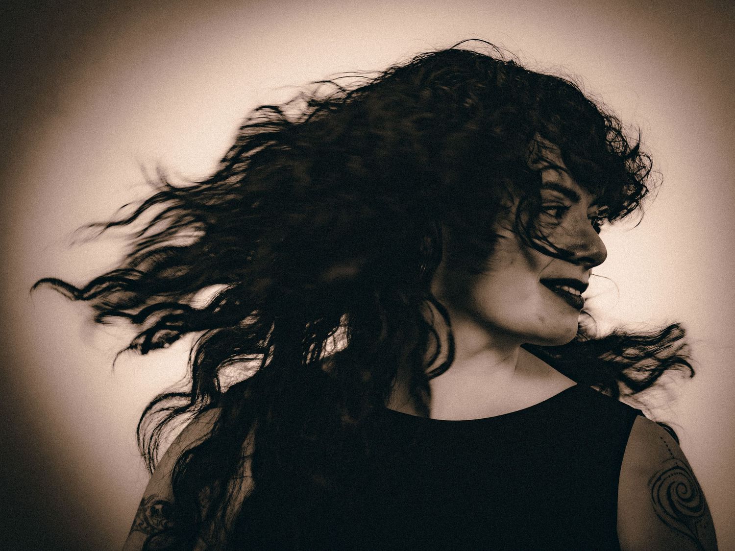 Dynamic sepia portrait of a woman with long curly hair blowing wildly, tattoo on shoulder.