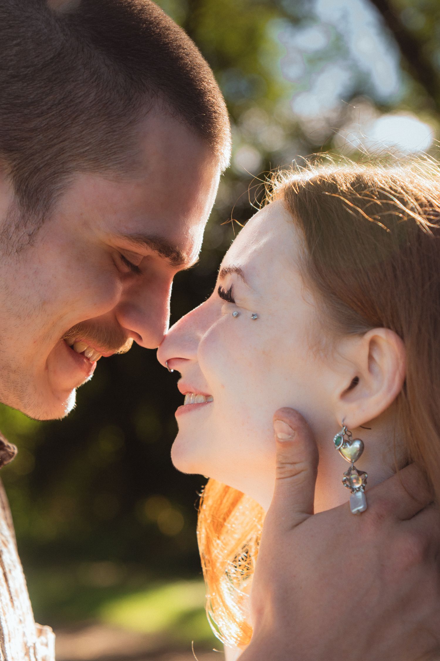 Romantic couple nearly kissing outdoors, woman with red hair and heart earrings, man gently holding her face in warm sunlight.