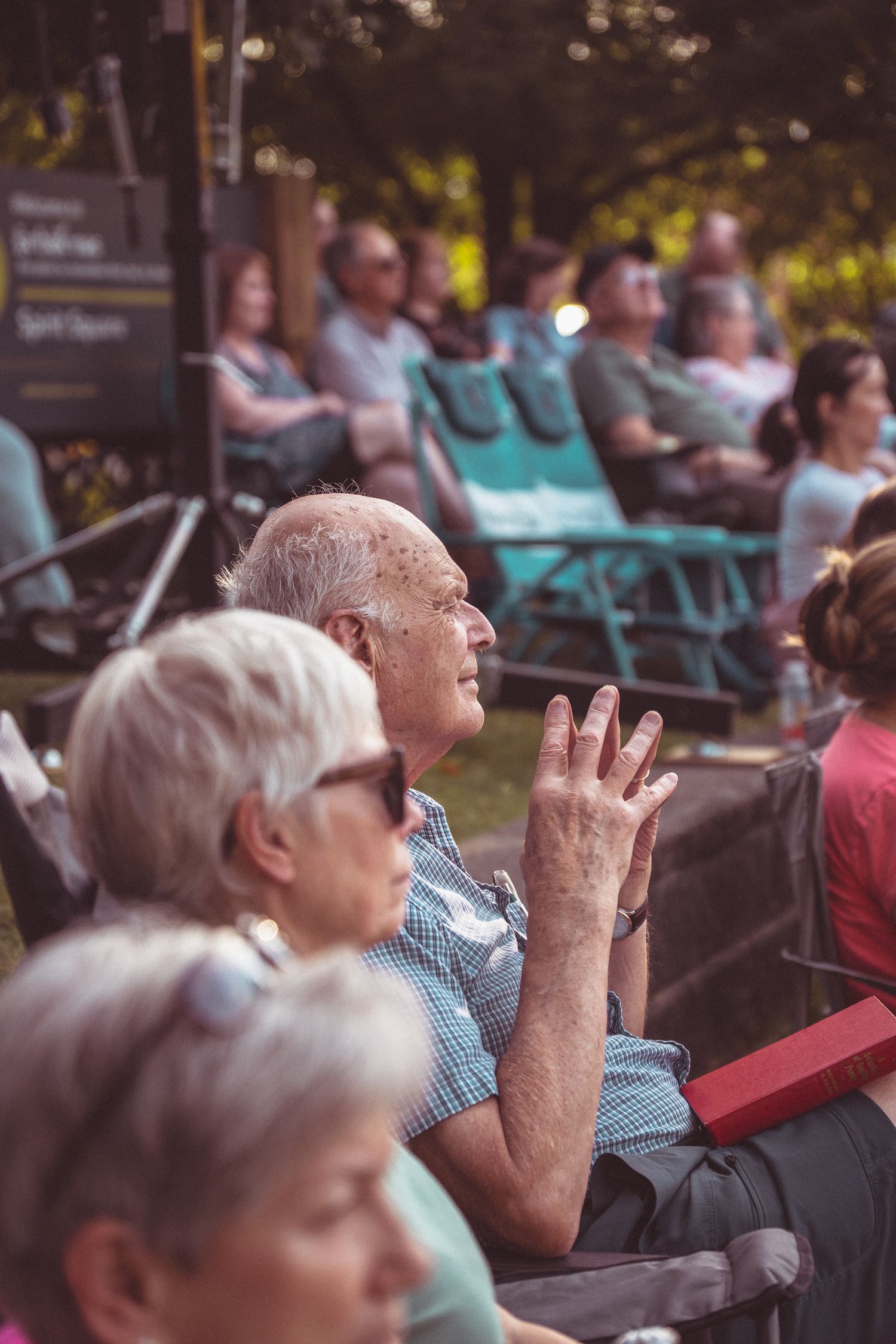 Elderly audience members sitting outdoors in chairs, attentively watching an open-air performance in a park setting.