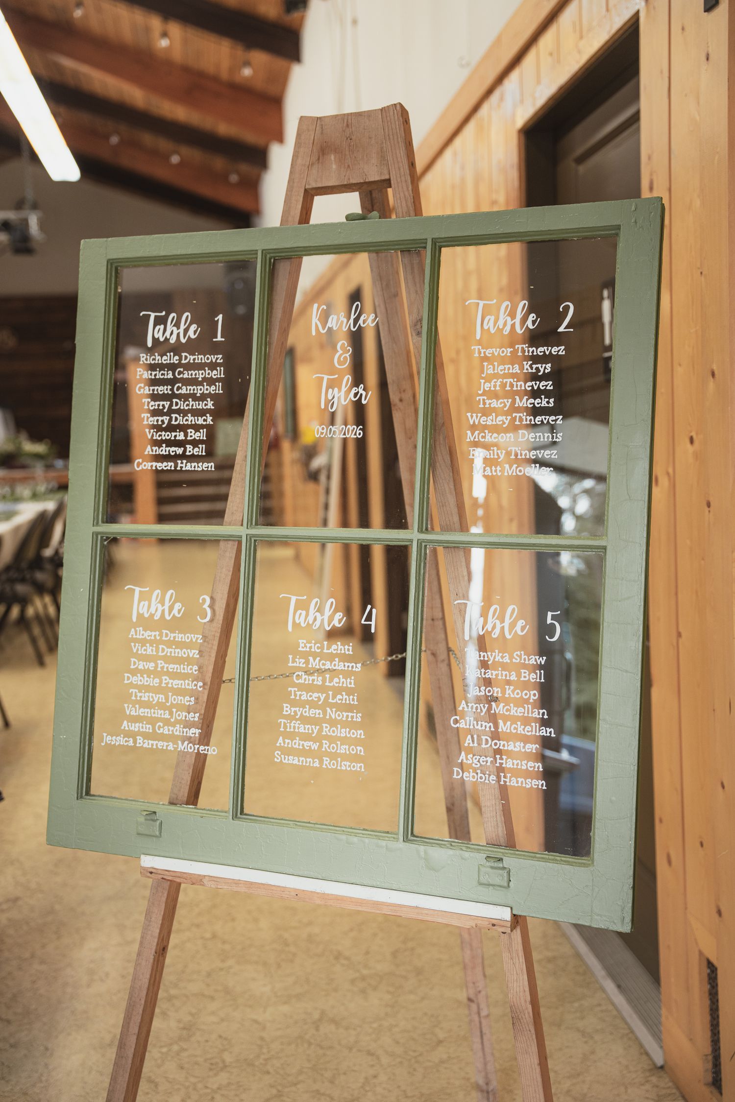 Rustic wedding seating chart displayed on vintage green window frame on wooden easel in barn venue.