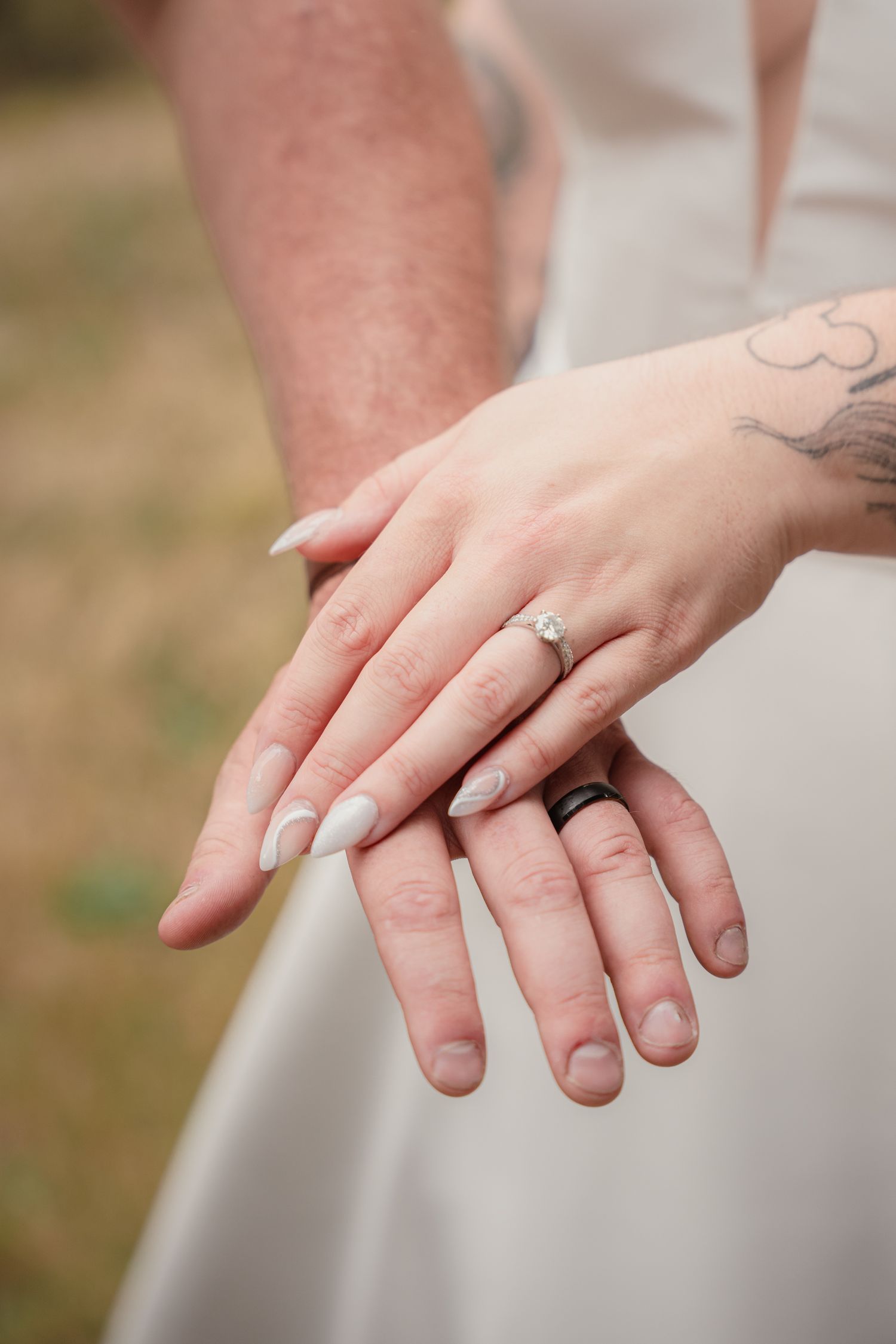 Bride and groom displaying wedding rings, bride wearing diamond ring with white nails, groom with black band, tattoo visible.
