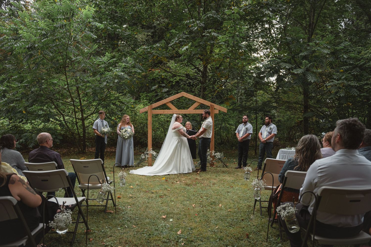 Outdoor woodland wedding ceremony with bride and groom at wooden arch altar, flanked by bridesmaids and groomsmen on lush green grass.