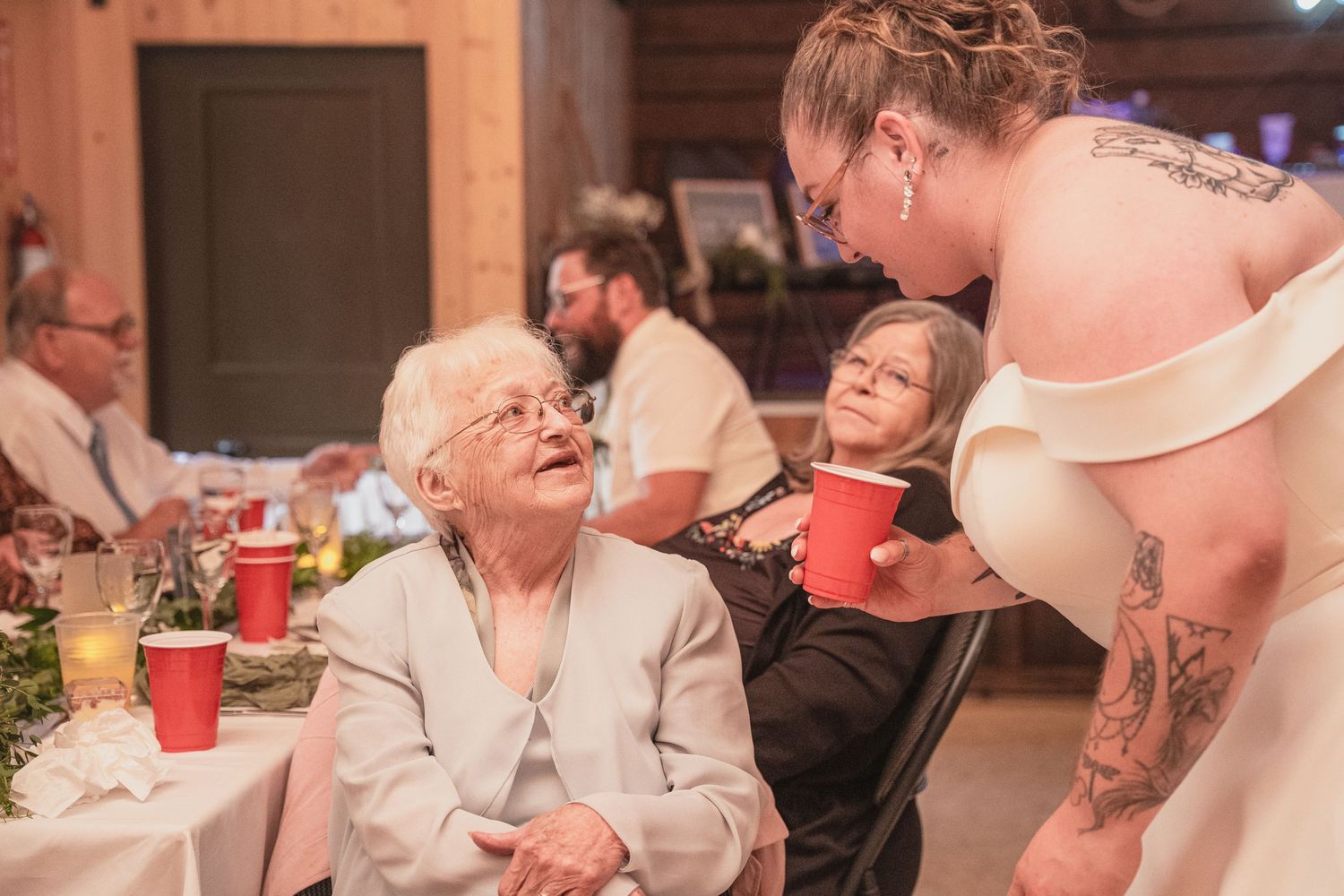 Tattooed bride in white gown lovingly greets elderly seated guest at wedding reception table.