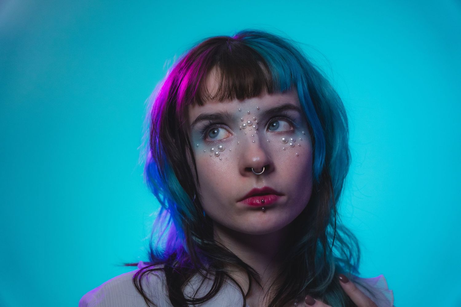 Woman with teal hair and pearl face gems gazing upward under neon blue and pink lighting.