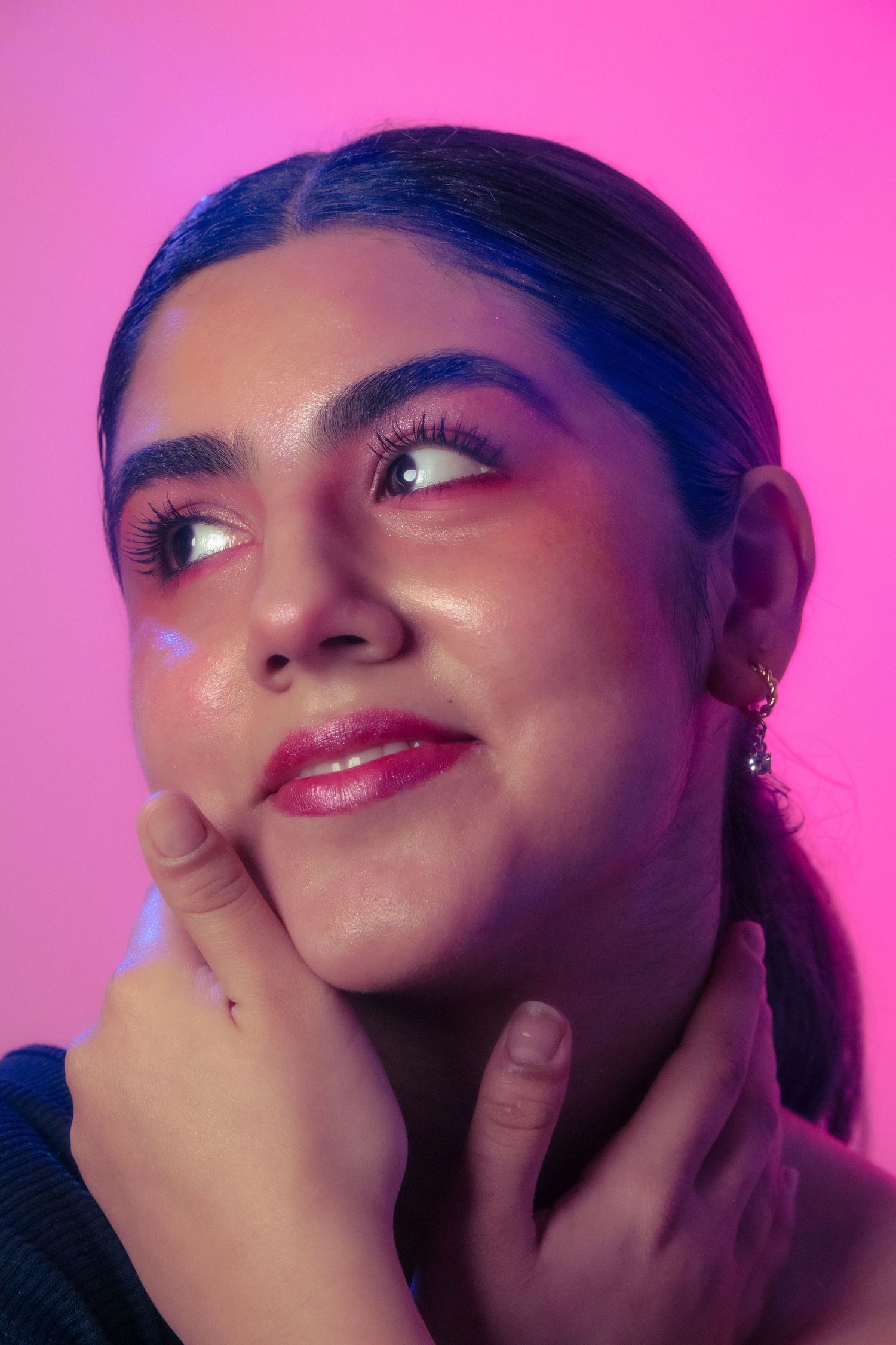A young woman with dramatic lashes and pink makeup smiles upward while touching her chin in neon pink light.