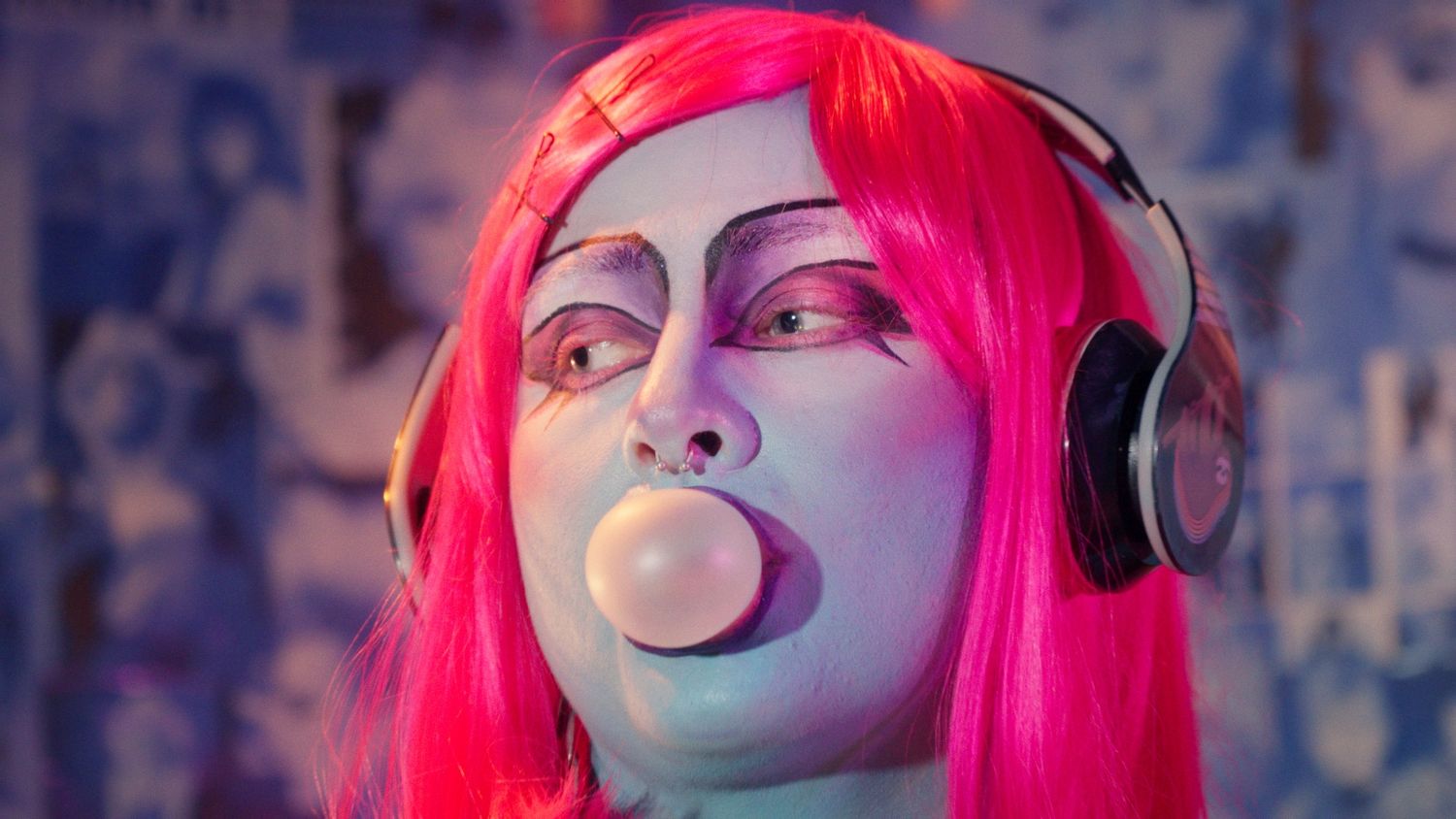 Person with pink hair and blue makeup blowing a bubble gum bubble while wearing headphones.