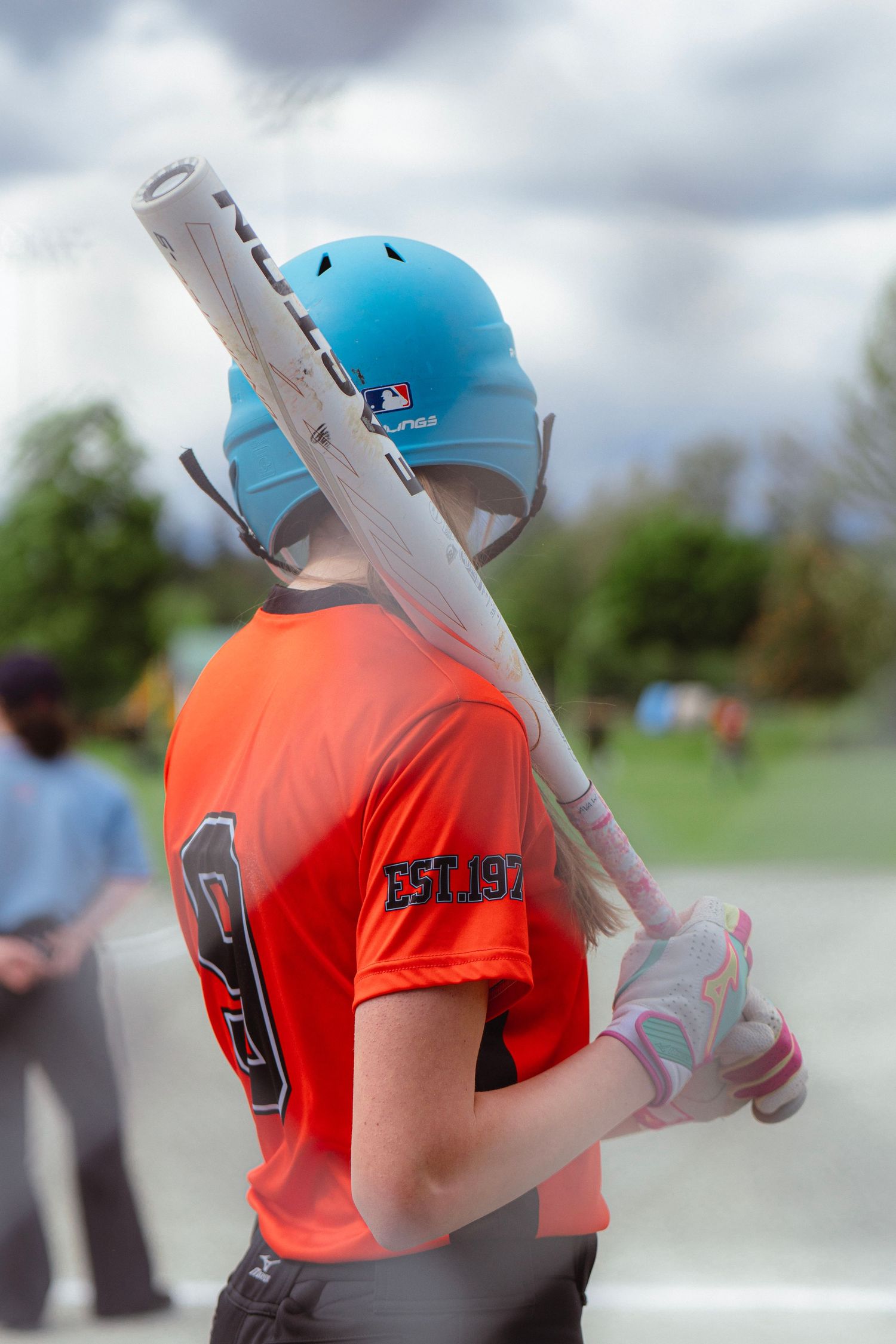 Female softball player in orange jersey and blue Rawlings helmet holds bat over shoulder at plate.