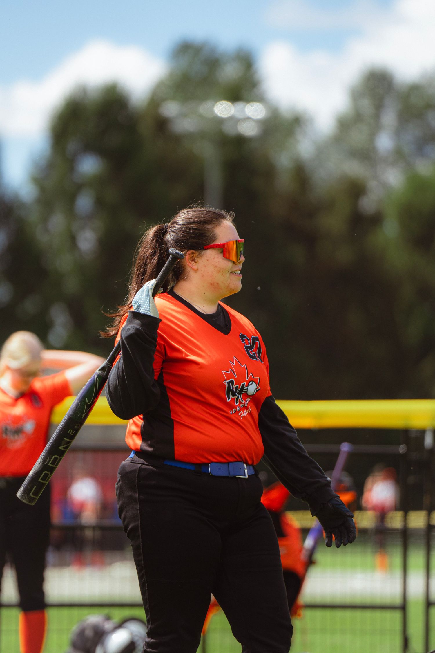 Female softball player in orange jersey resting bat on shoulder outdoors.
