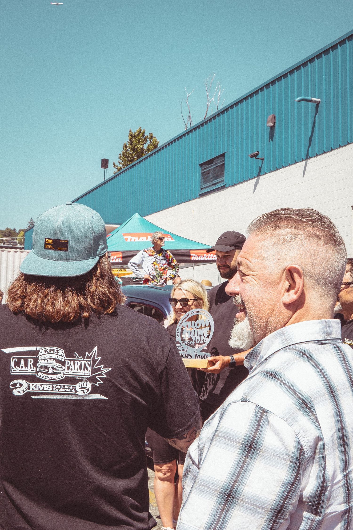 Smiling man in plaid shirt at outdoor car show, CAB Parts KMS logo shirt in foreground.