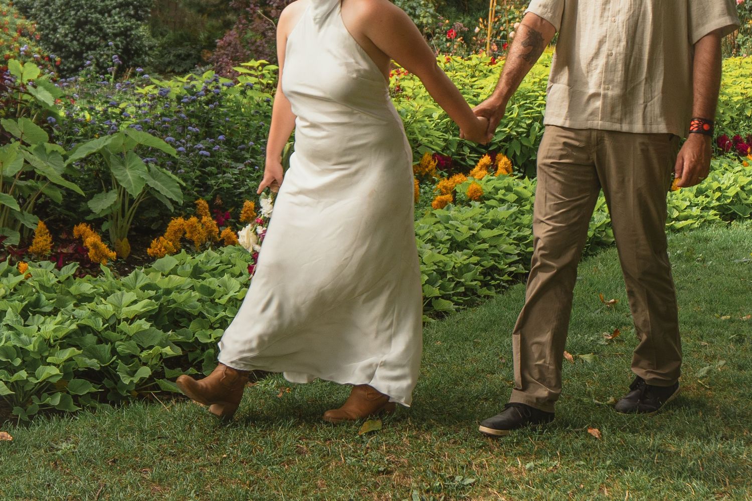 Couple holding hands walking through a lush garden, woman in white dress and man in khaki pants.