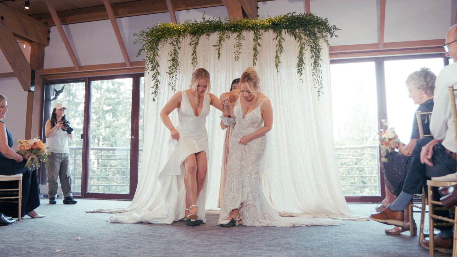 Two brides in white gowns share a tender moment at their wedding ceremony beneath a floral arch.