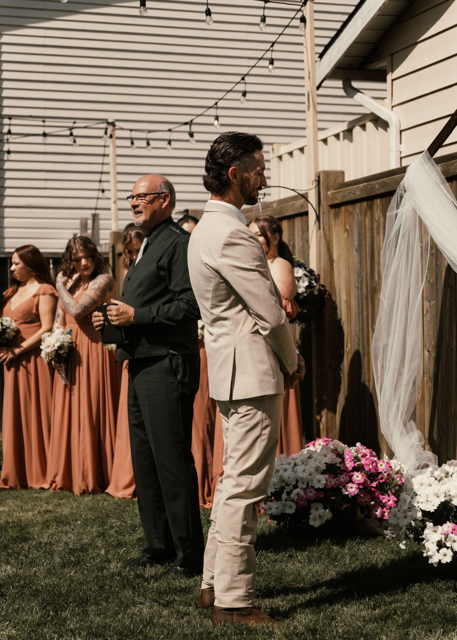 Groom in tan suit awaits bride at outdoor backyard wedding ceremony with bridesmaids in rust-colored gowns nearby.