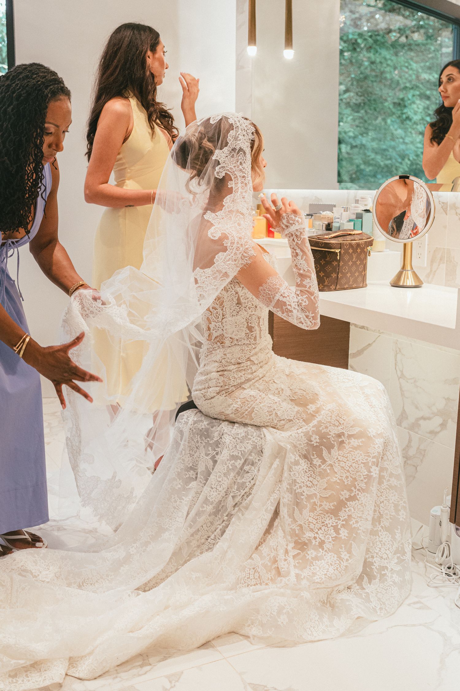 Bride in lace wedding gown and veil being helped by bridesmaids in a elegant bridal suite before wedding ceremony.