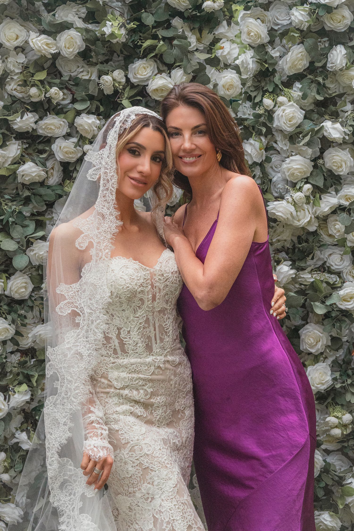 Bride in lace gown and veil poses with woman in purple dress before white rose floral wall backdrop.