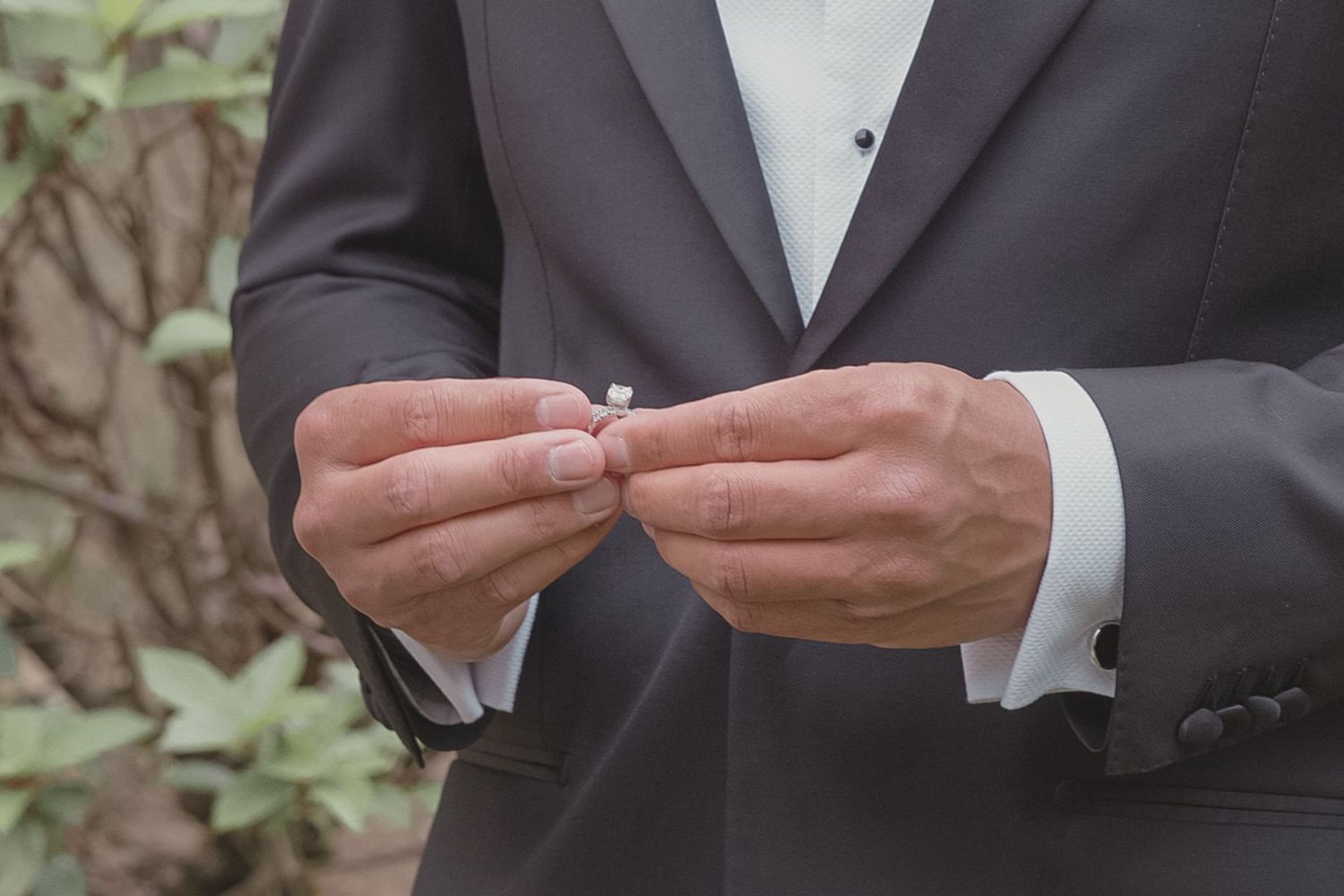 Man in black suit holding a diamond engagement ring between his fingers outdoors.