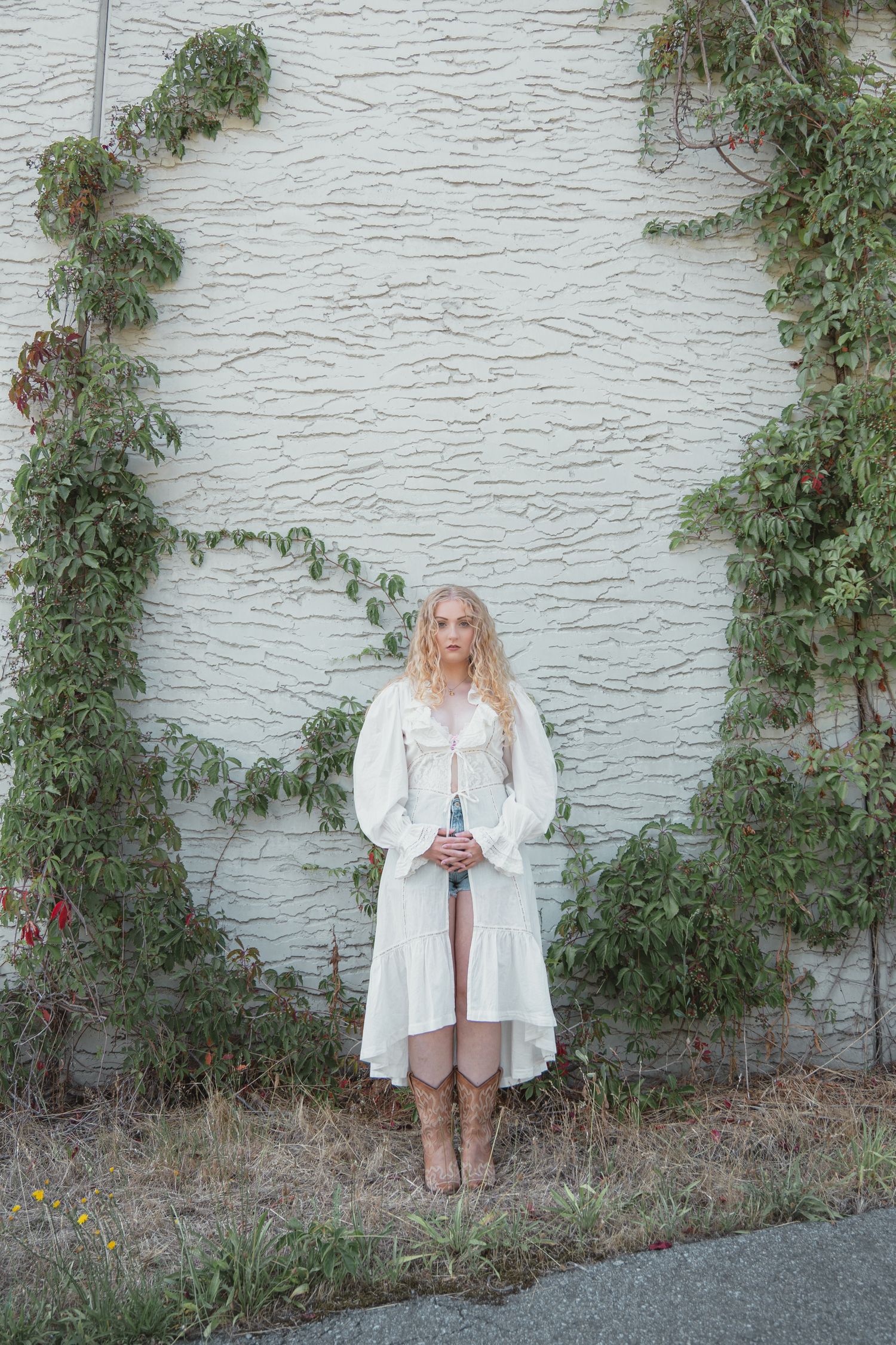 Young blonde woman in white dress and cowboy boots posing against ivy-covered textured wall.