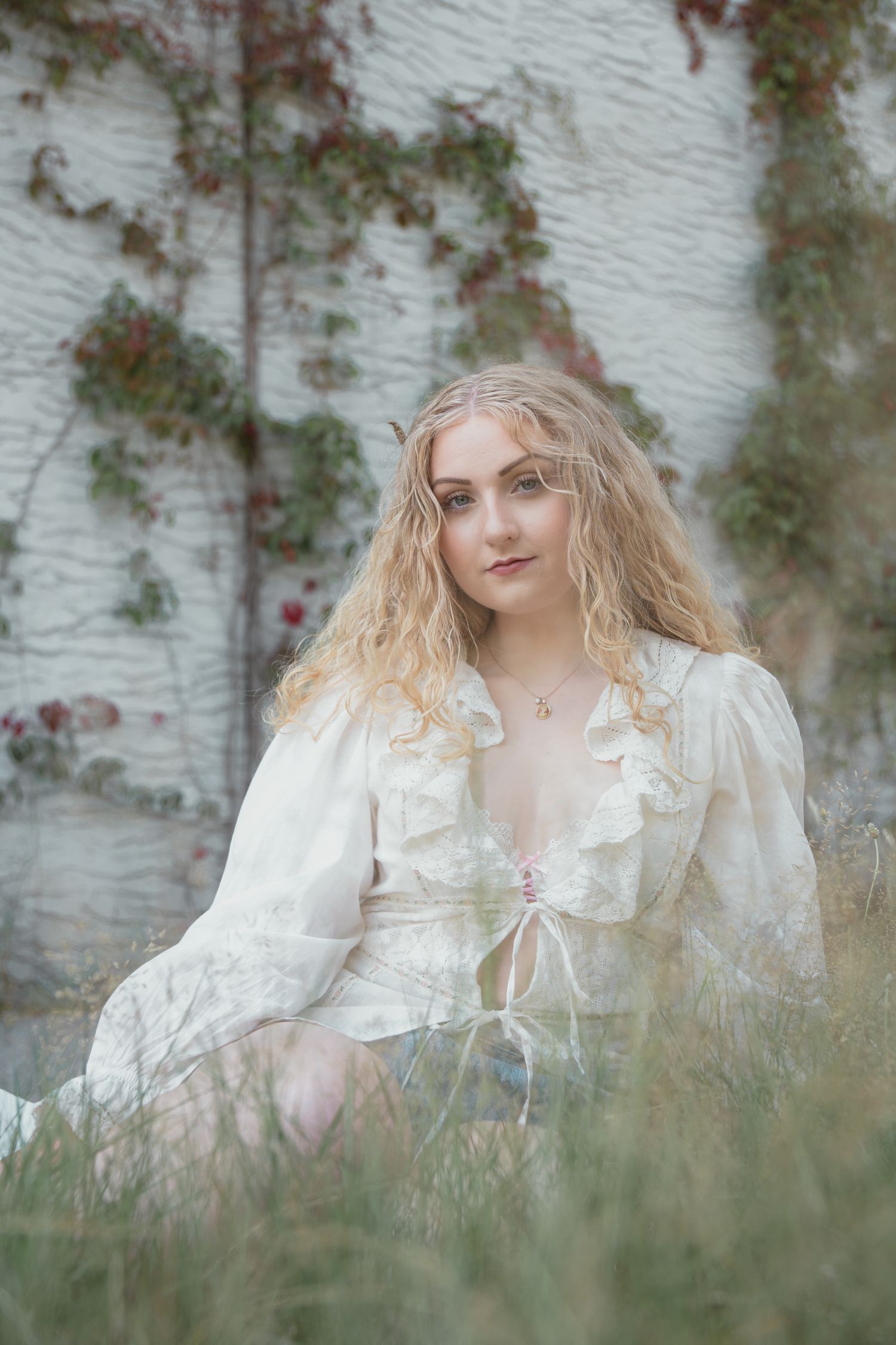 Young woman with curly blonde hair sits in grass wearing a white ruffled dress near an ivy-covered white brick wall.
