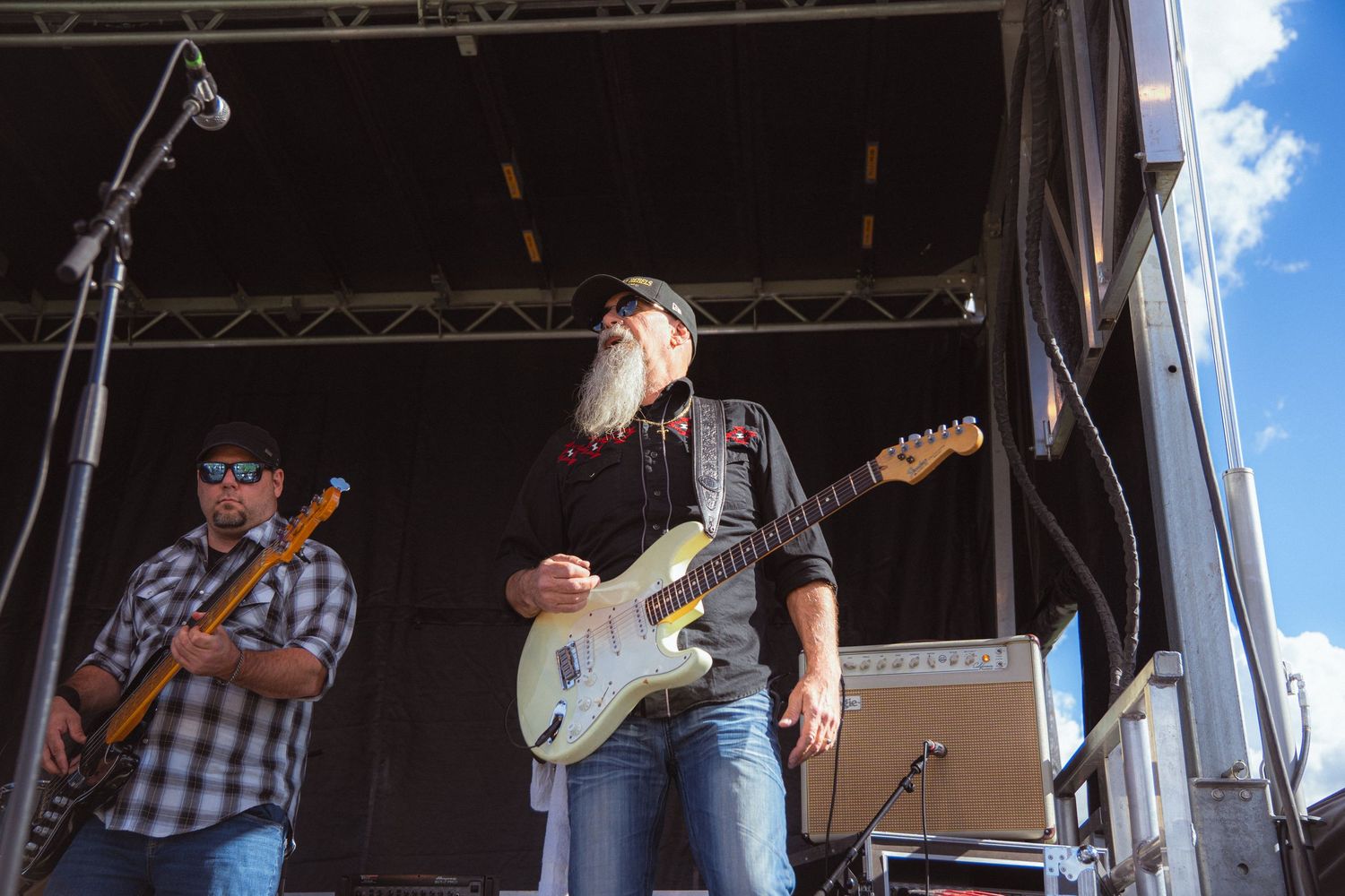 Two guitarists performing on outdoor festival stage with amplifier under clear sky.