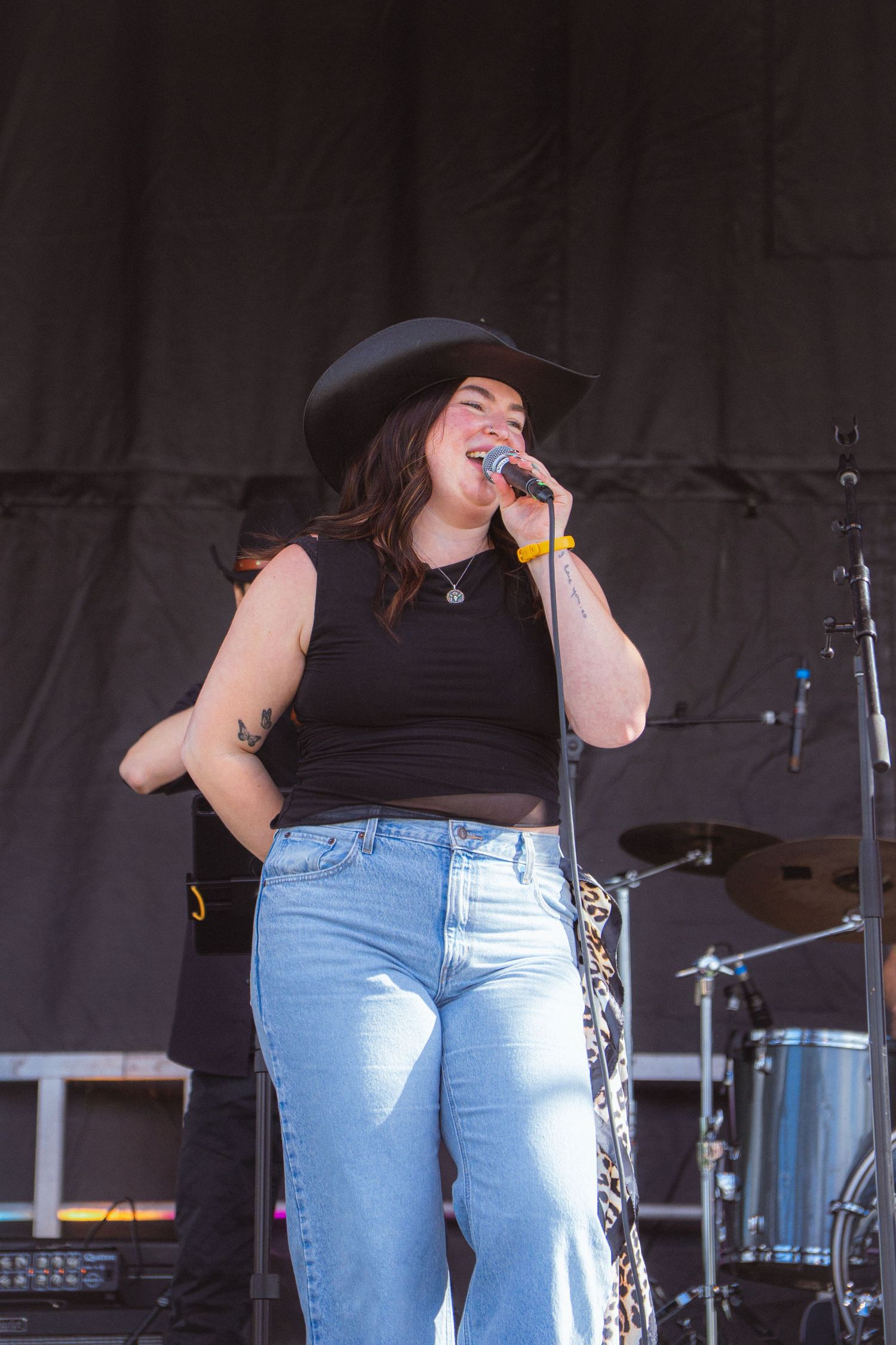 Female singer in black cowboy hat and jeans performs with microphone on outdoor festival stage.