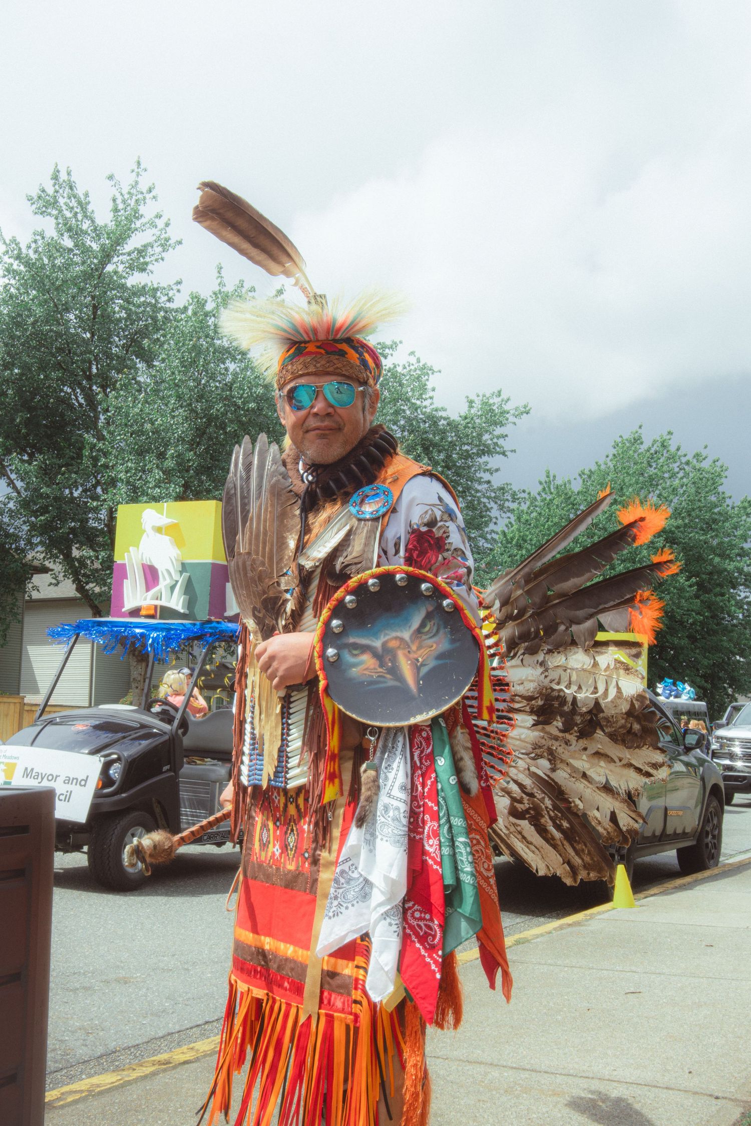 Indigenous dancer in full traditional regalia with feathered headdress holds a drum at a street parade.