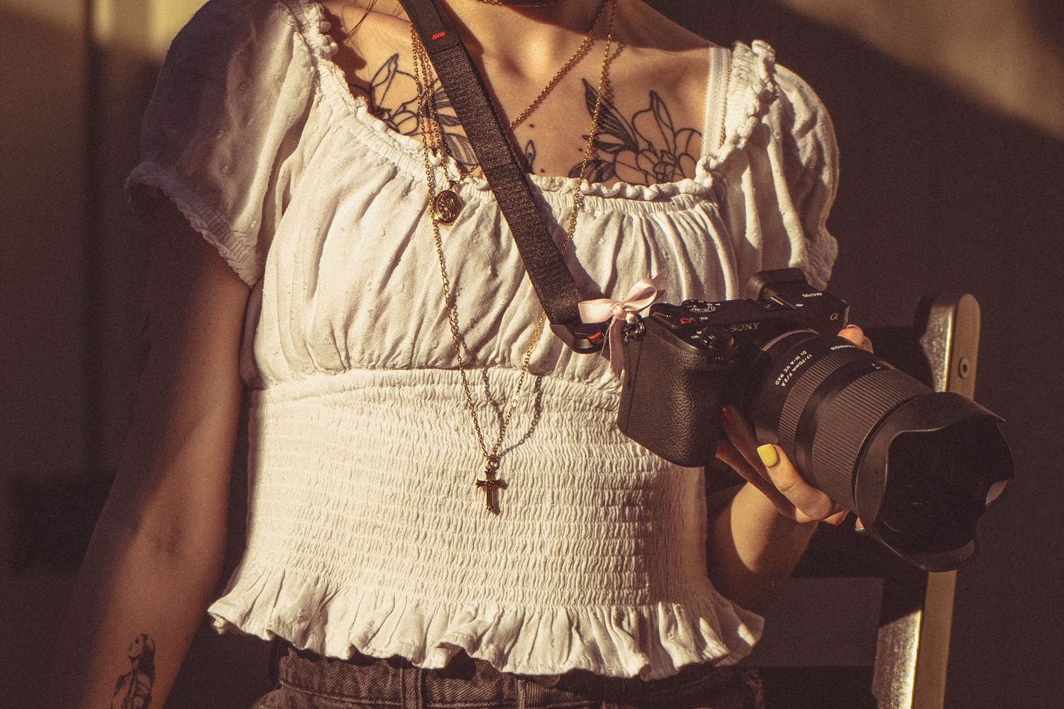 Woman in white smocked crop top holds DSLR camera with cross necklace and butterfly tattoo visible.