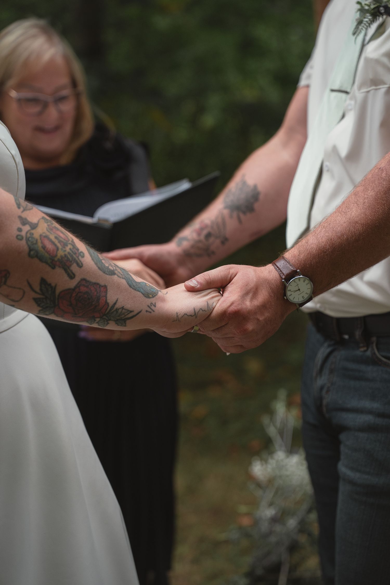 Tattooed bride and groom hold hands during vows as officiant reads from book at outdoor wedding.