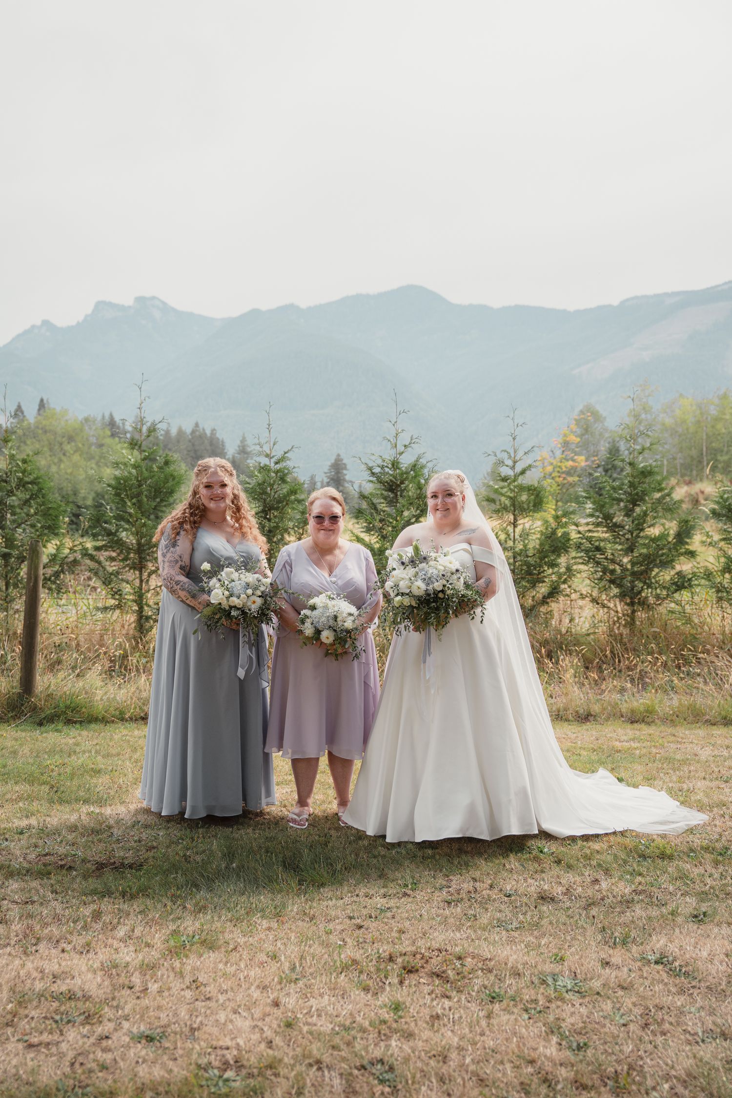 Bride in white gown with two bridesmaids holding bouquets outdoors with mountain backdrop.