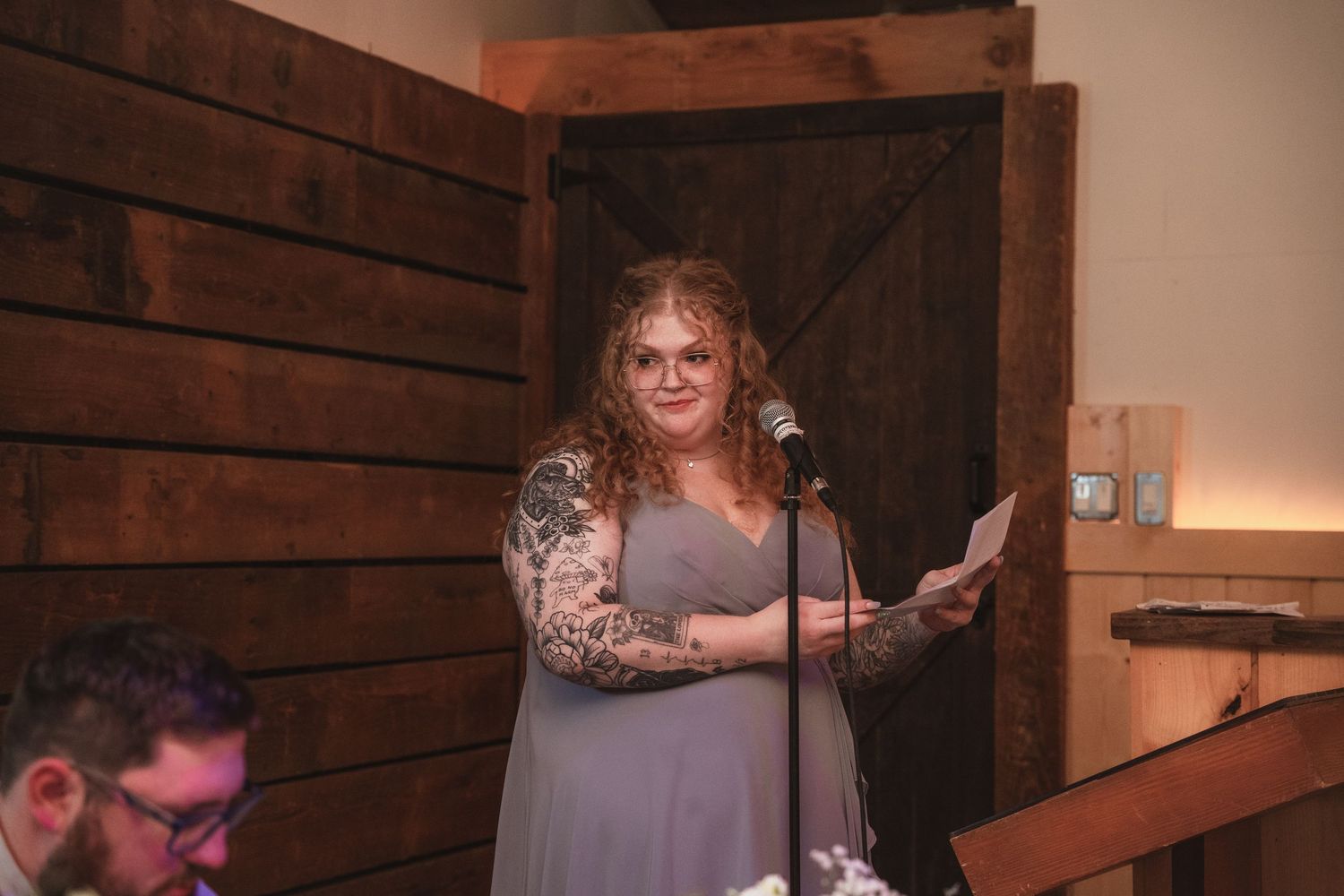 Tattooed bridesmaid in gray dress reads heartfelt speech at microphone during rustic barn wedding ceremony.