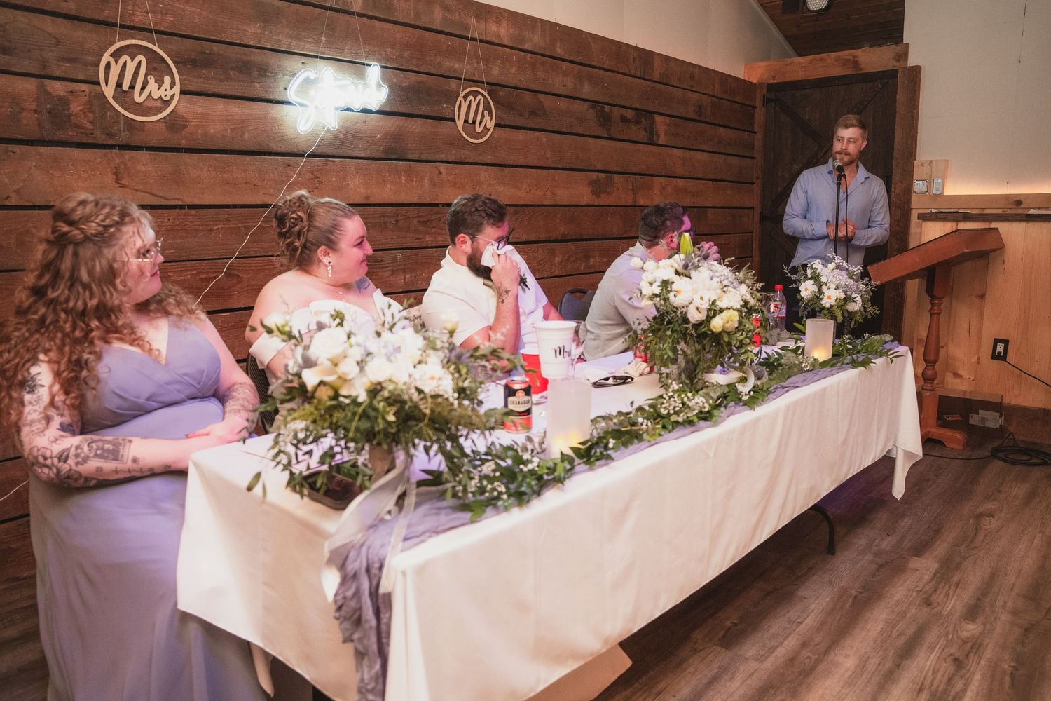 Wedding party seated at head table while groomsman delivers speech at rustic barn reception.