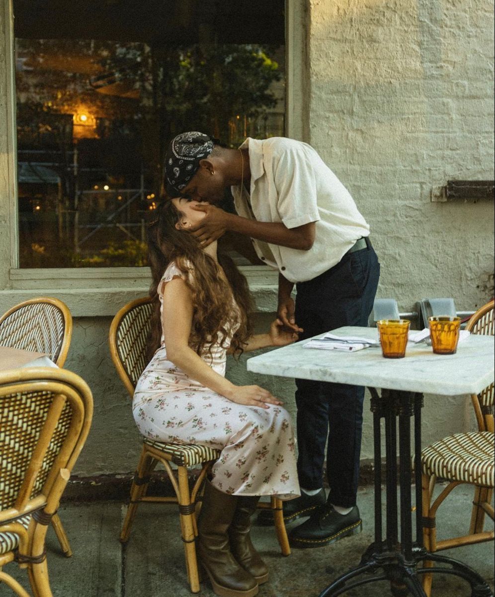 A couple shares a romantic kiss at an outdoor cafe patio table with amber drinks at dusk.