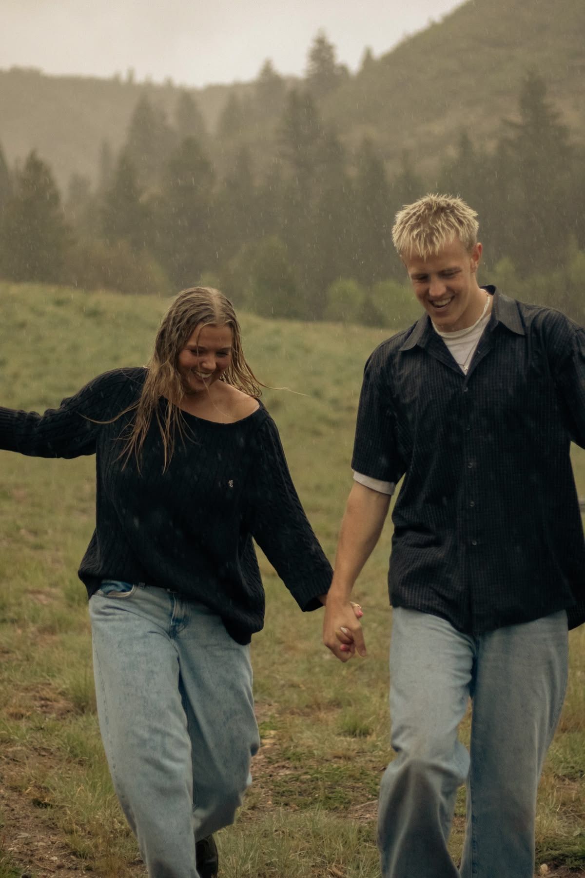 A young couple holds hands walking in the rain on a green hillside with misty mountains behind them.