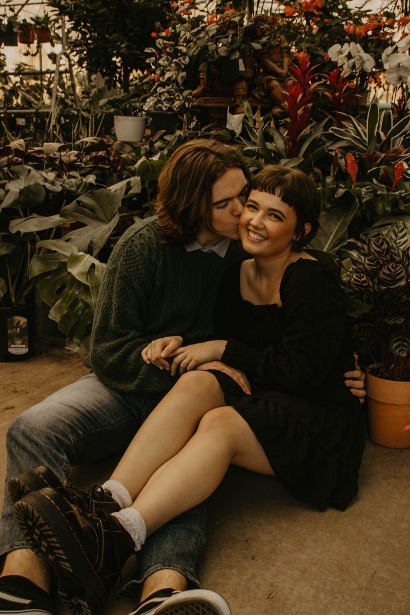 A couple sits on the floor of a lush greenhouse, sharing a tender kiss among vibrant tropical plants.