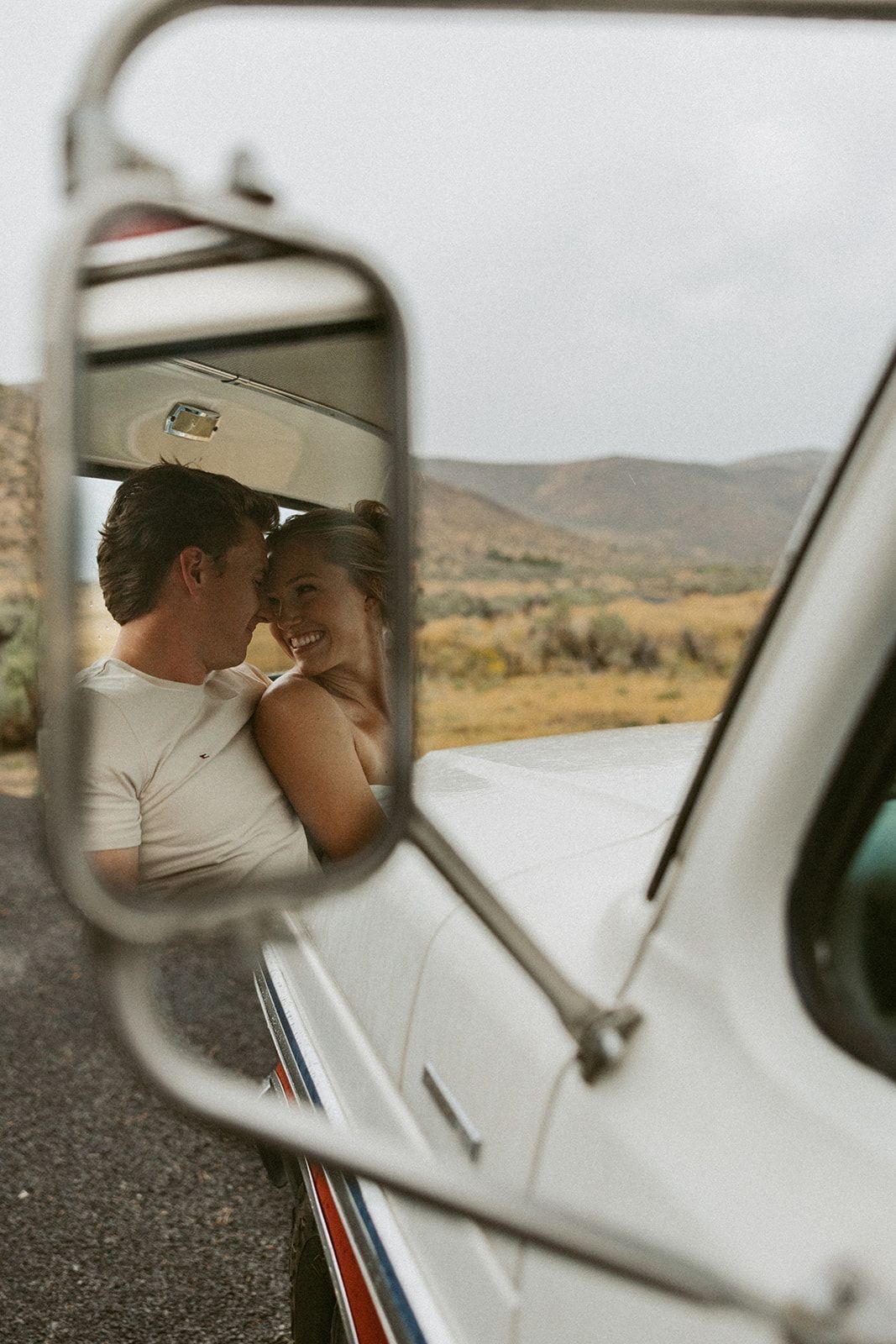Couple embracing and smiling at each other reflected in a vintage white van side mirror with mountains behind.