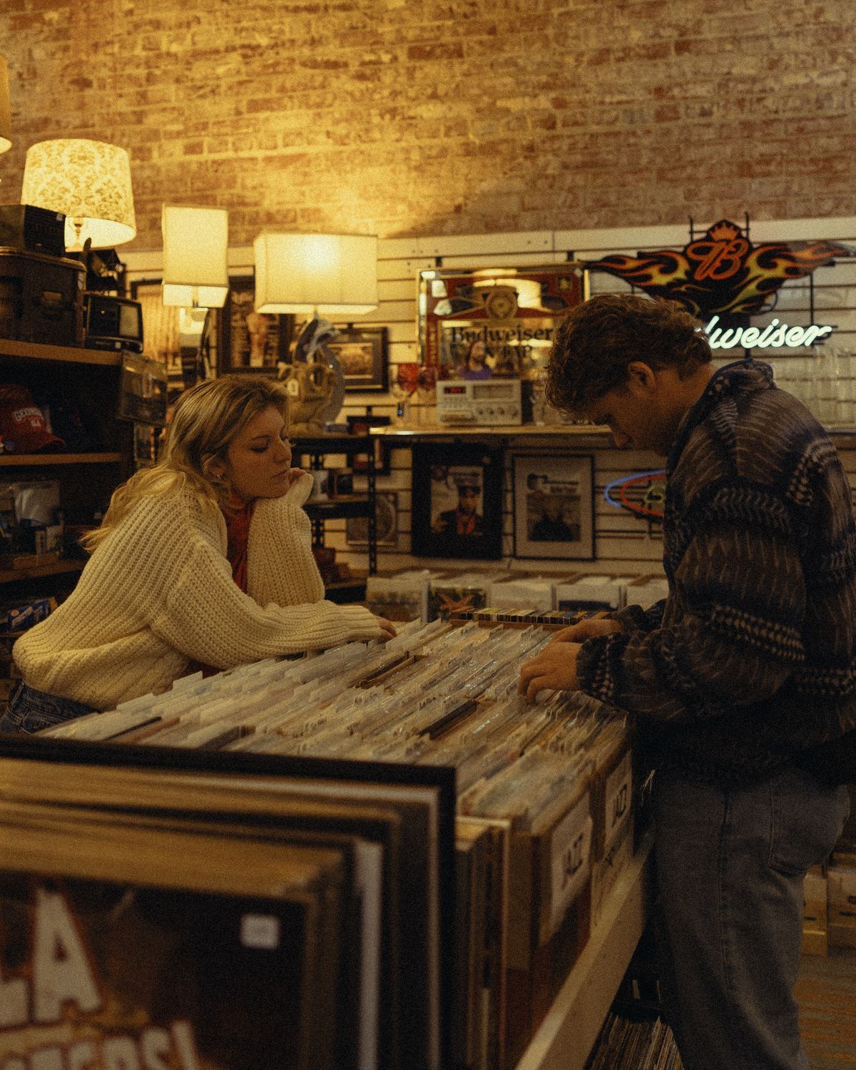 Two people browsing vinyl records in a cozy, warmly lit record store with brick walls and a Budweiser neon sign.